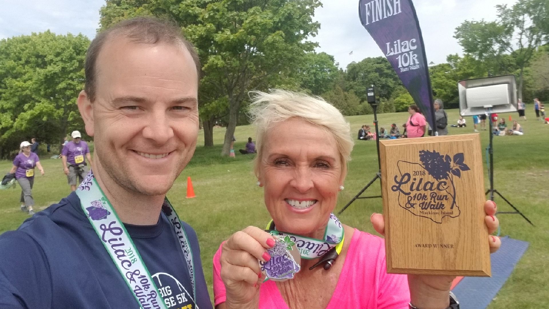 Selfie of a smiling man in navy and a woman in pink at the park finish line of the Lilac 10k Run/Walk. She holds a wooden Award Winner plaque; purple finish banner and onlookers in the background.