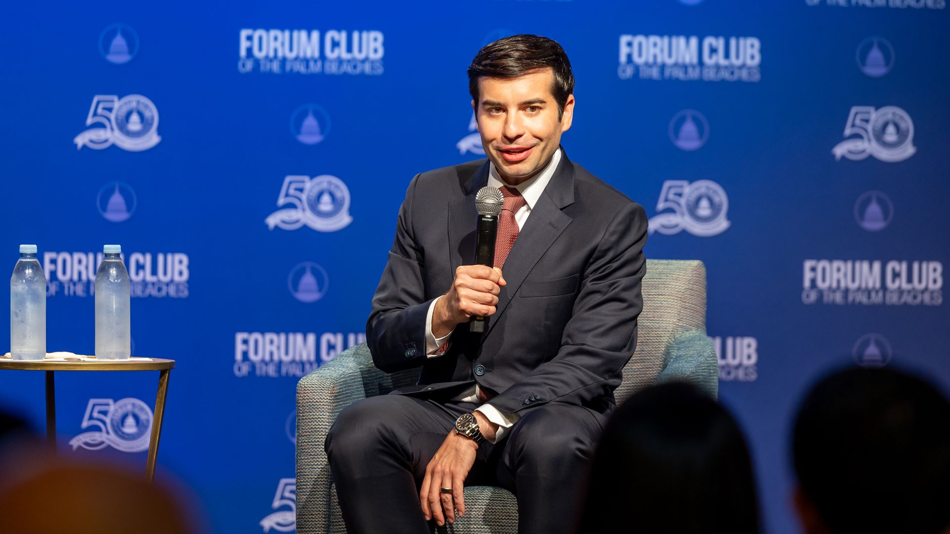 Keith Sonderling in a dark suit and red tie sits on a stage, holding a microphone. He is in front of a blue backdrop with Forum Club of the Palm Beaches logos, and two water bottles on a table in foreground.
