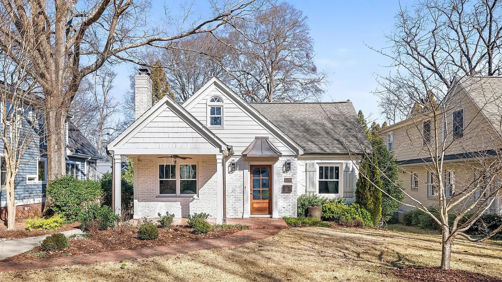 White brick house with gray roof, wooden front door, and a covered porch surrounded by leafless trees and brown grass lawn under clear blue sky.