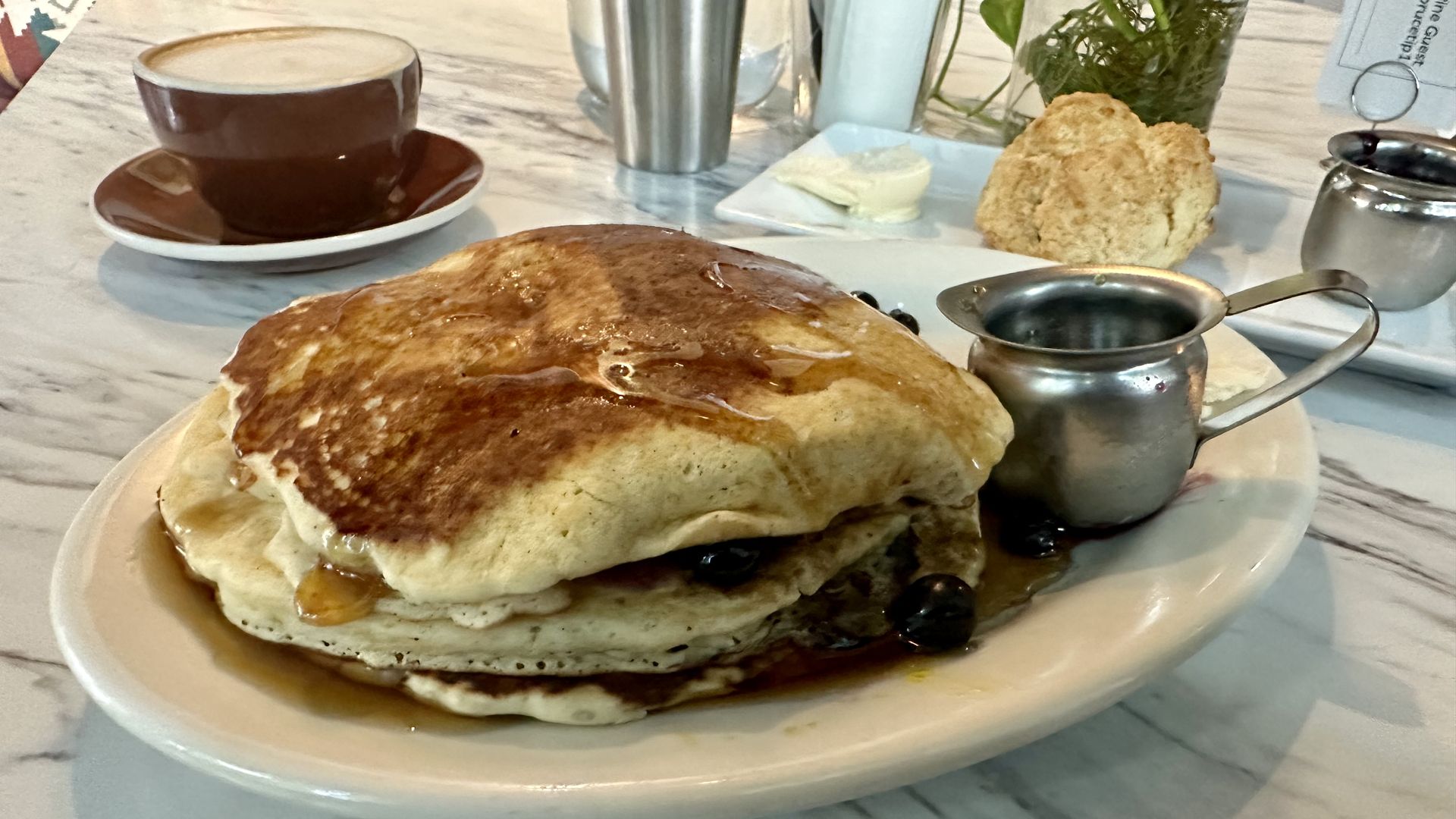 A stack of blueberry pancakes with syrup running down the sides, on a marble toned table with a cappuccino and biscuit visible in the background.