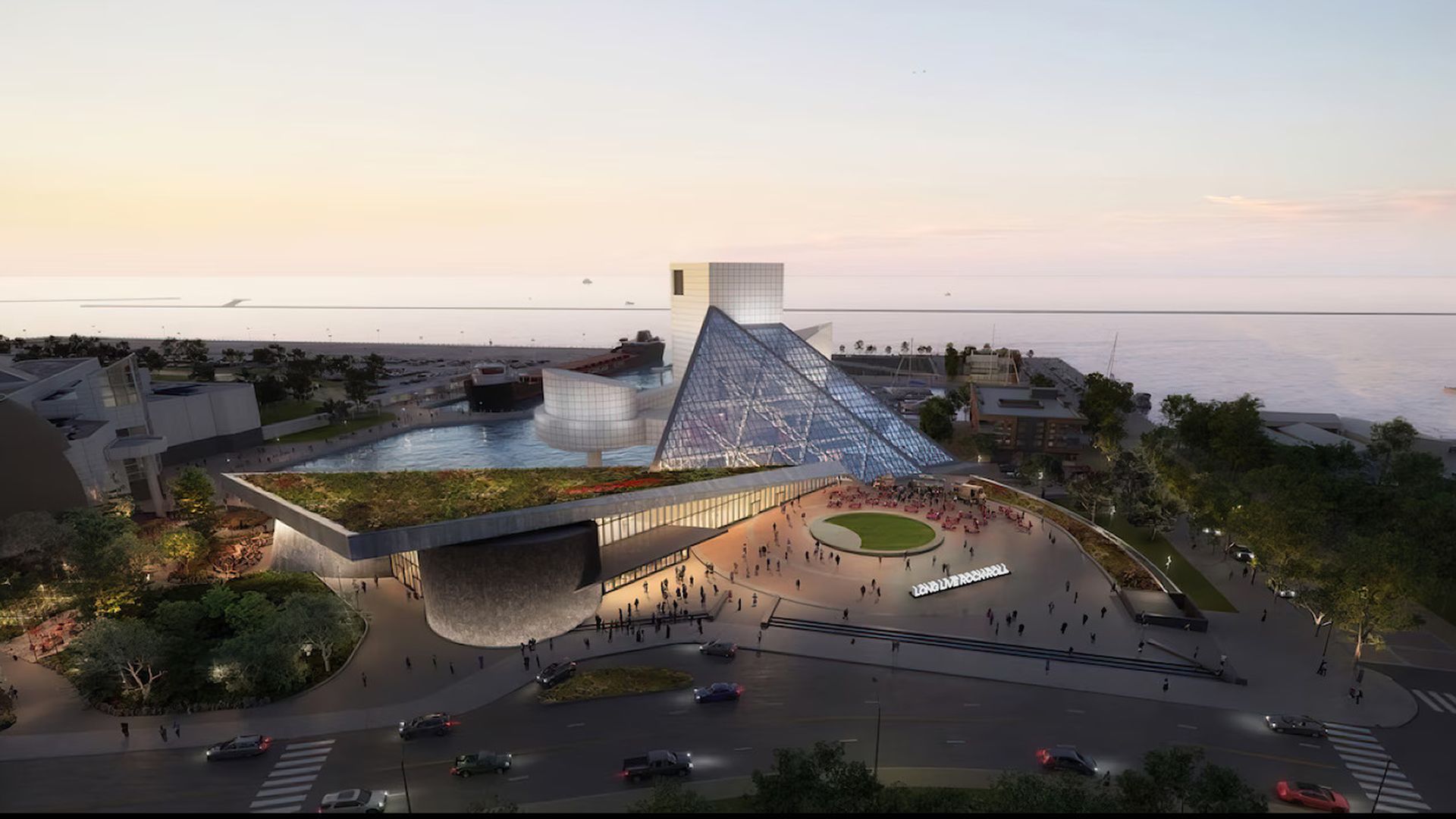 Futuristic waterfront plaza at sunset dominated by a glass pyramid, a curved building extension, and a circular grassy plaza with people strolling along the promenade by the water.