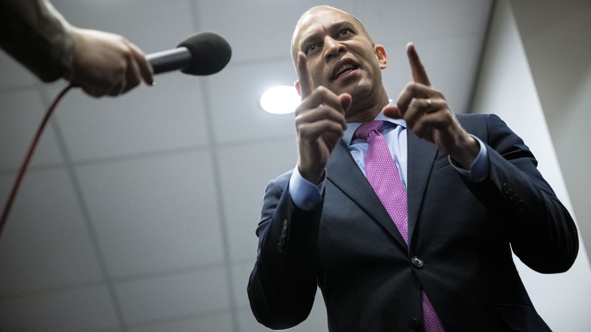 House Minority Leader Hakeem Jeffries, wearing a gray suit, blue shirt and purple tie, speaking into a microphone under a white ceiling.