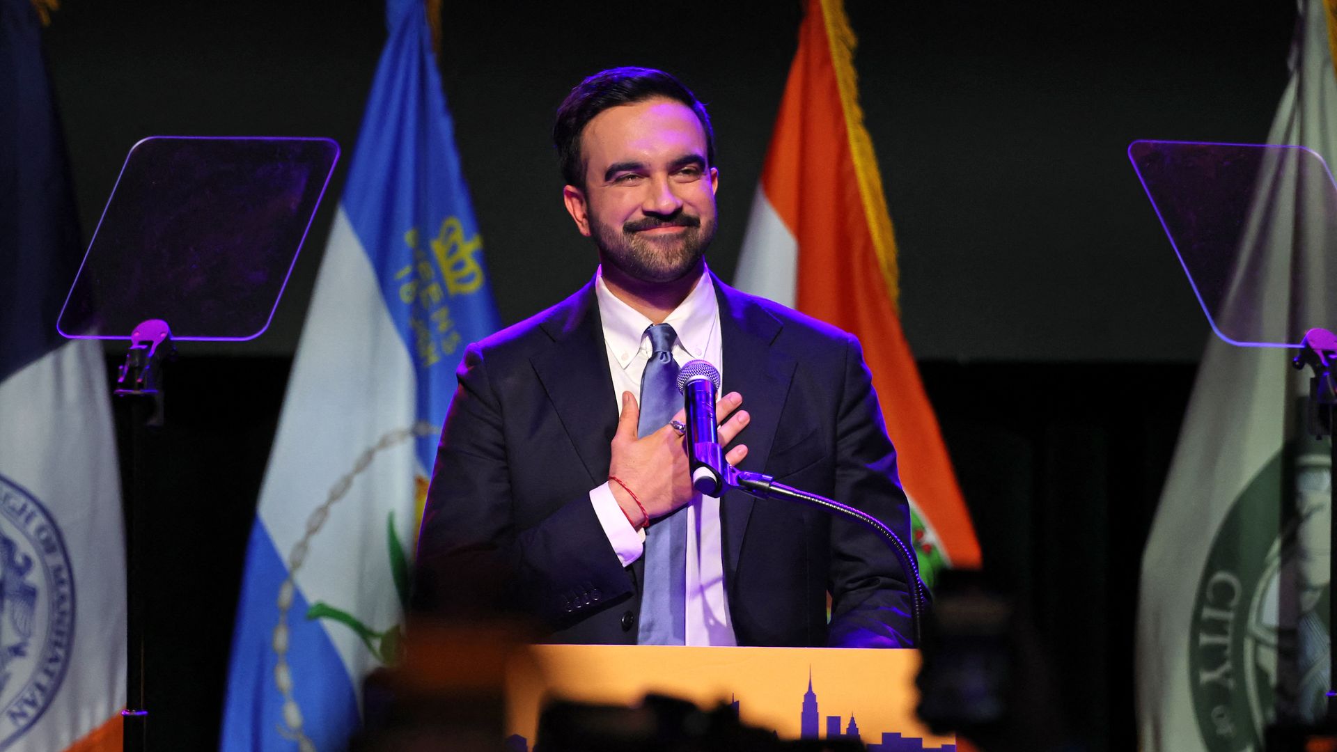 Zohran Mamdani holds a hand to his chest and smiles behind a podium, with a row of flags behind him. 