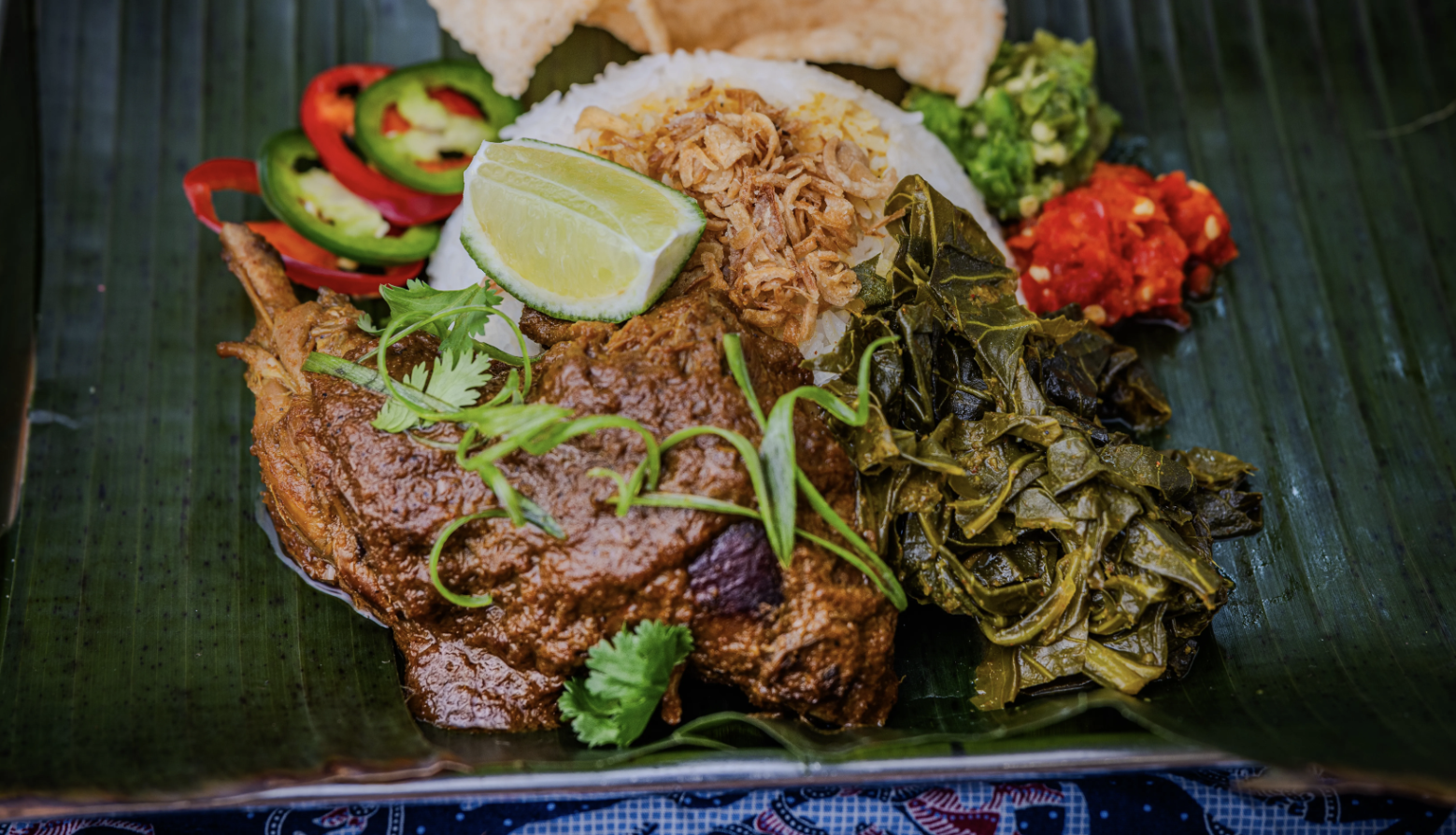 Plate on banana leaf with brown spiced chicken, white rice topped with fried onions and lime, cooked greens, sliced peppers, green salsa, red chili sauce, and crackers.
