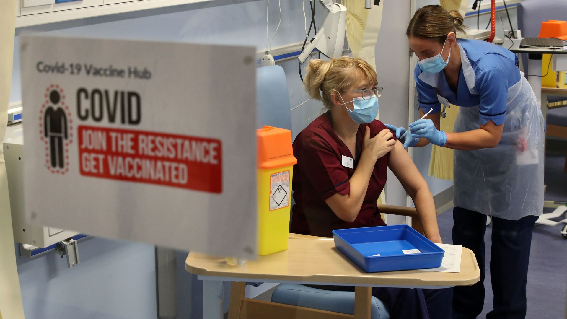 A woman wearing a face mask gives a shot to another woman wearing a face mask, who sits at a desk behind a sign that says "COVID: Join the resistance get vaccinated"