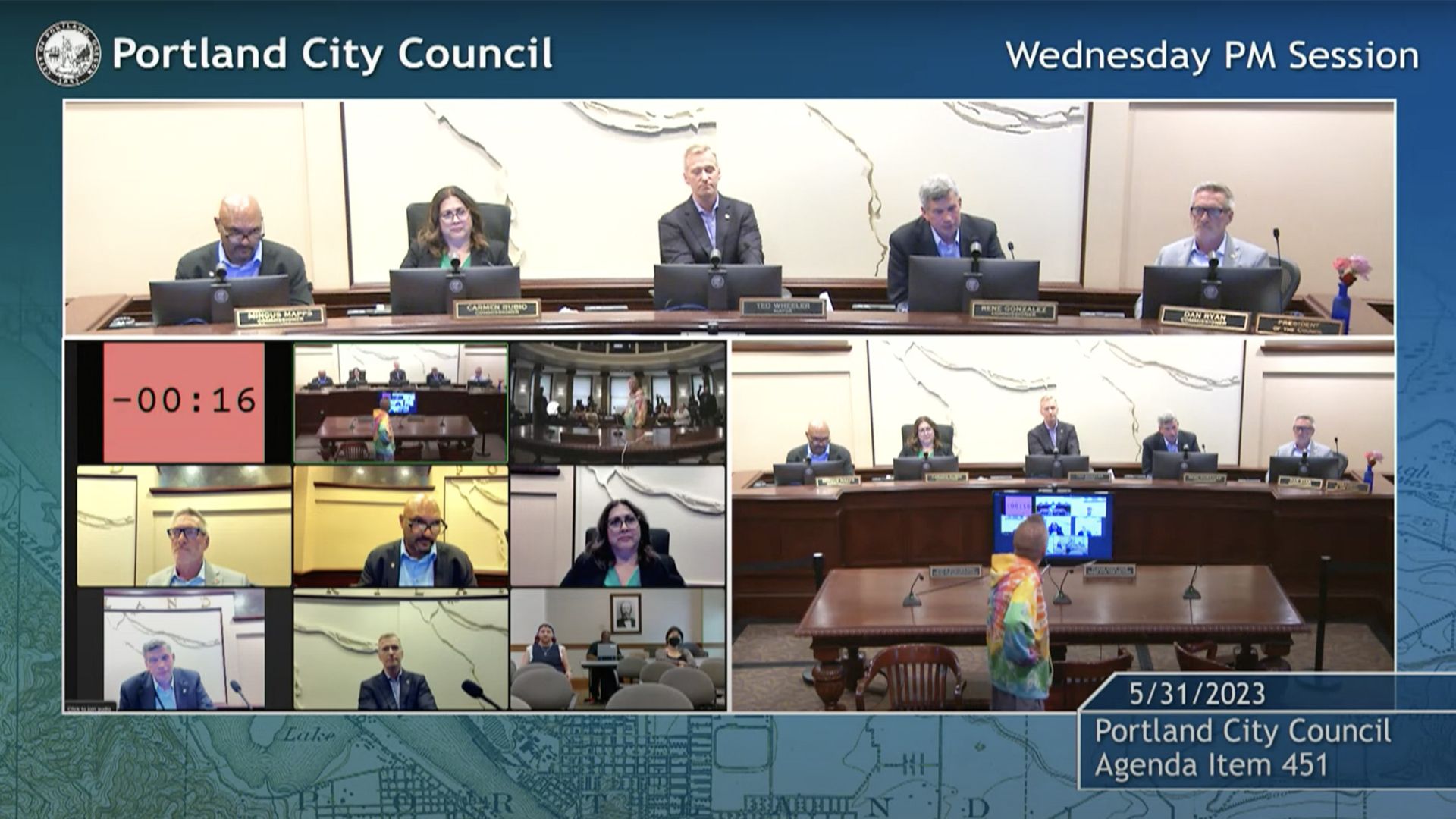 A screenshot of the broadcast of public meeting with elected officials on a dais in the back and a man in a tie-dyed t-shirt standing and addressing them with one hand on his hip. A red countdown clock shows the speaker has run out of time.