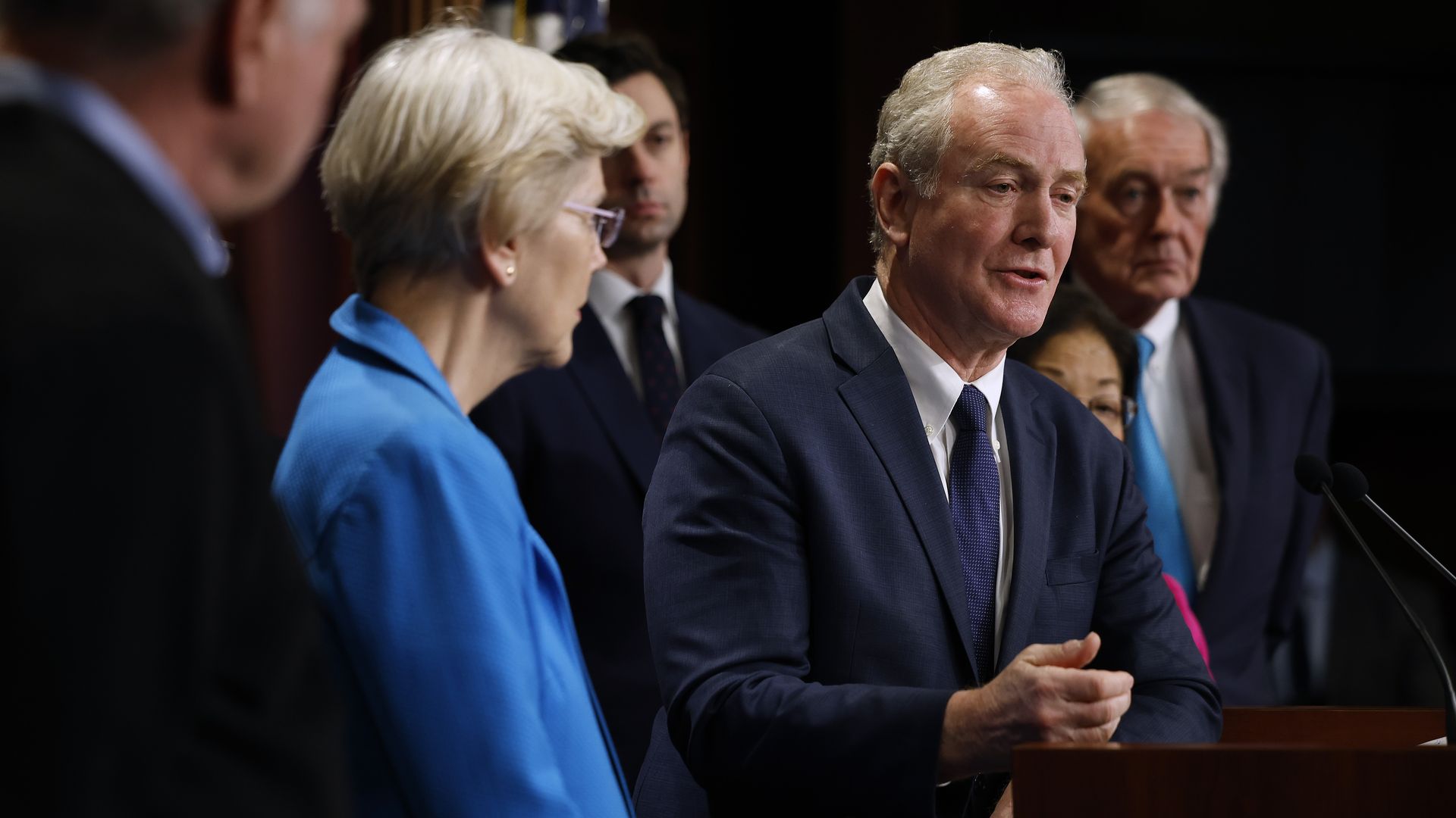 Sen. Chris Van Hollen, wearing a blue suit, white shirt and blue tie, speaking at a podium surrounded by Senate colleagues.