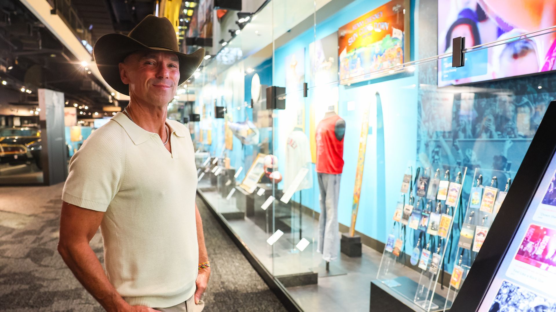 A smiling Kenny Chesney in a beige polo shirt and cowboy hat stands beside a glass storefront display with blue walls, mannequins wearing outfits, and bright posters overhead.
