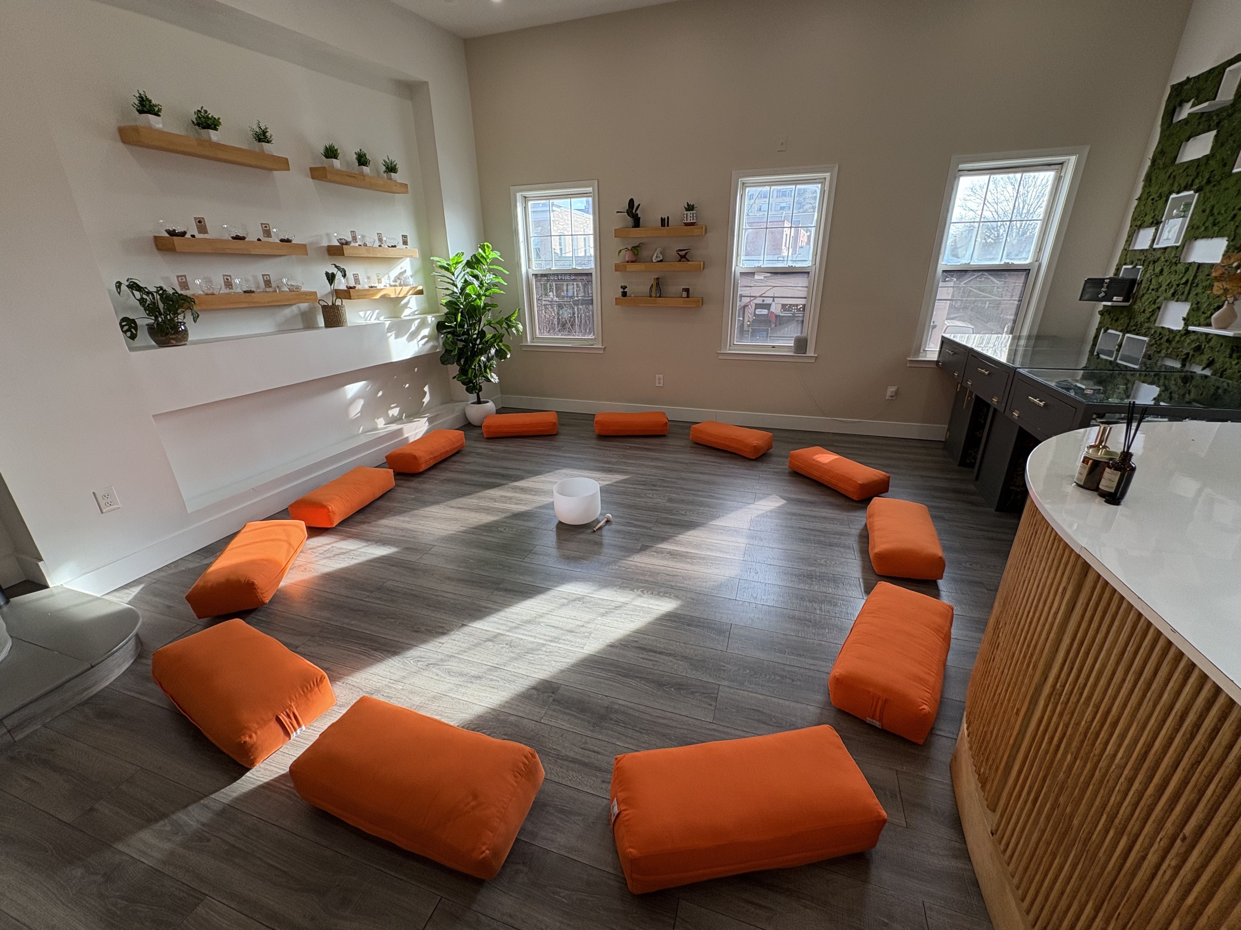 Bright modern room with gray wood floor, orange cushions placed on the floor, white shelves with small plants along left wall, and a green moss wall with frames on the right; large windows.