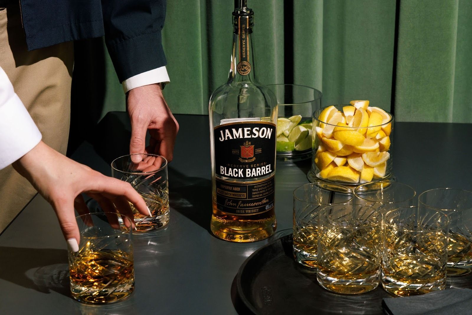 Jameson Black Barrel whiskey bottle on a dark table, surrounded by crystal glasses. A hand reaches for a glass as bowls of lemon wedges and lime slices sit nearby against a green curtain.