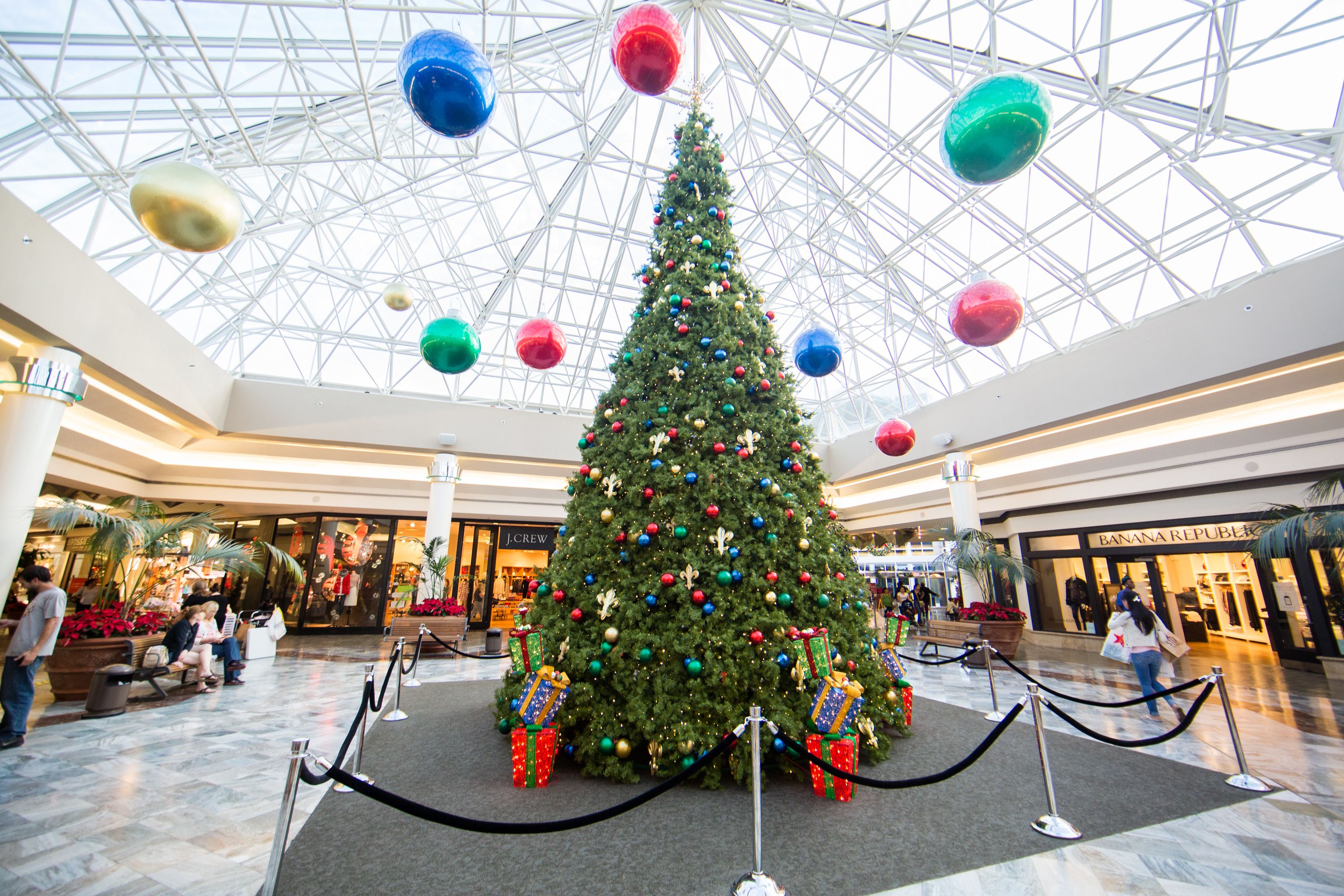 A large decorated Christmas tree sits underneath a glass atrium in the middle of a large shopping mall corridor.