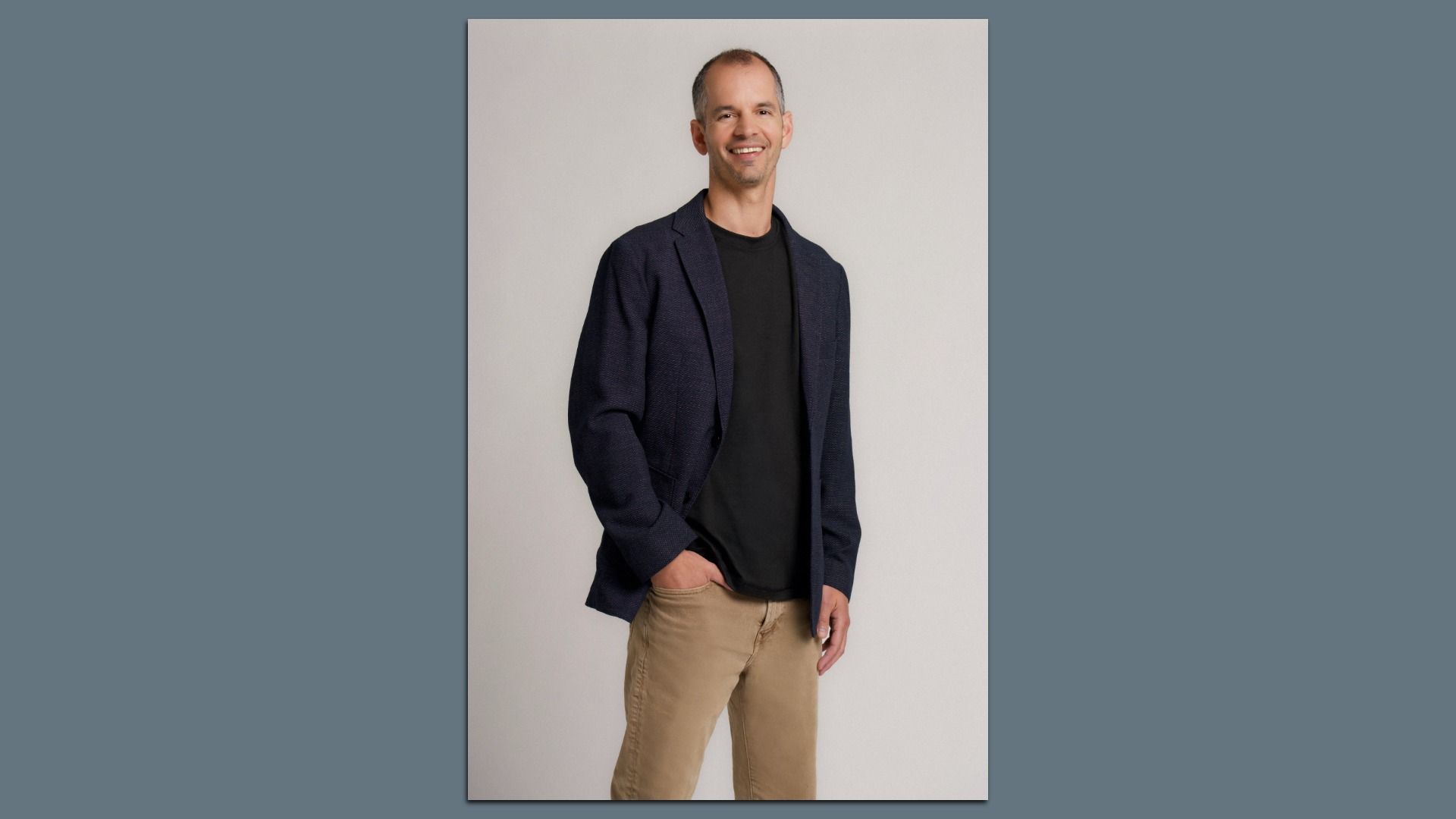 Smiling man wearing a navy blazer, black shirt, and tan khaki pants stands against a light gray backdrop with one hand in his pocket.