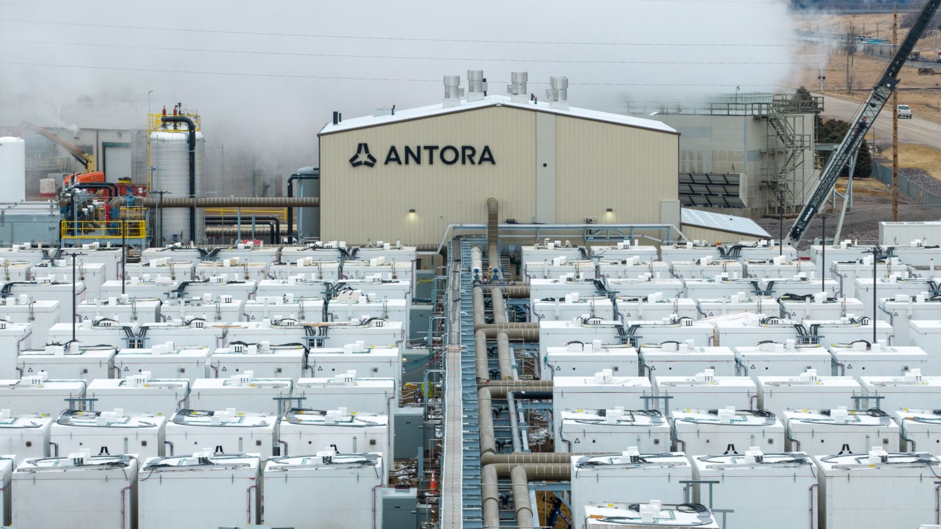Industrial energy facility with a beige building labeled "ANTORA" at center, surrounded by rows of white battery cabinets, extensive pipes, and smoke plumes in the background.