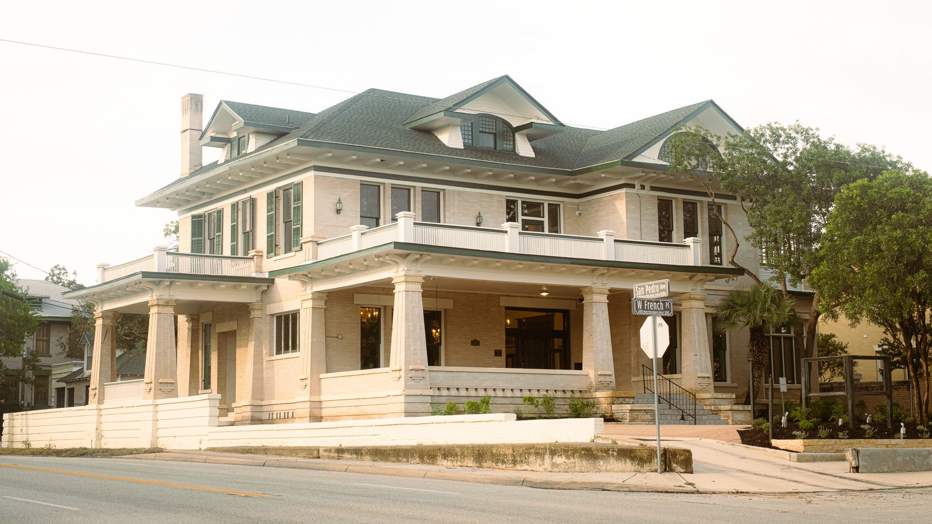 Large beige brick house with a green gabled roof, white railings and columns on a corner lot; street signs read San Pedro Ave and W French Pl, with trees on the right.
