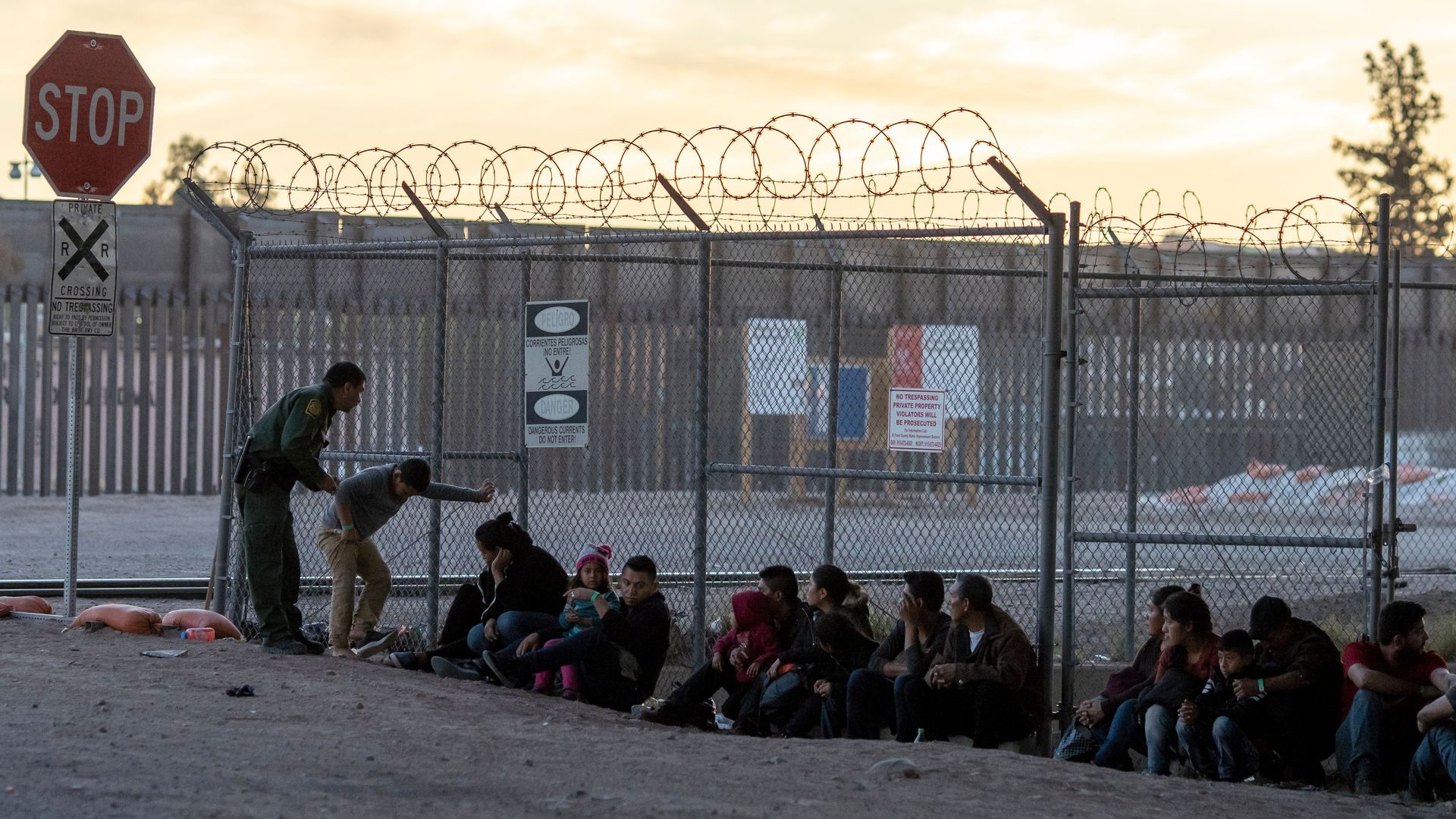 Migrants sitting against a chained link fence while one border patrol officer speaks to them. 
