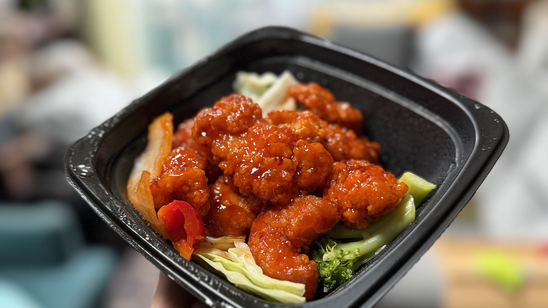 Hand holding a black takeout container with glazed orange chicken pieces over steamed broccoli, cabbage, and bell pepper slices.