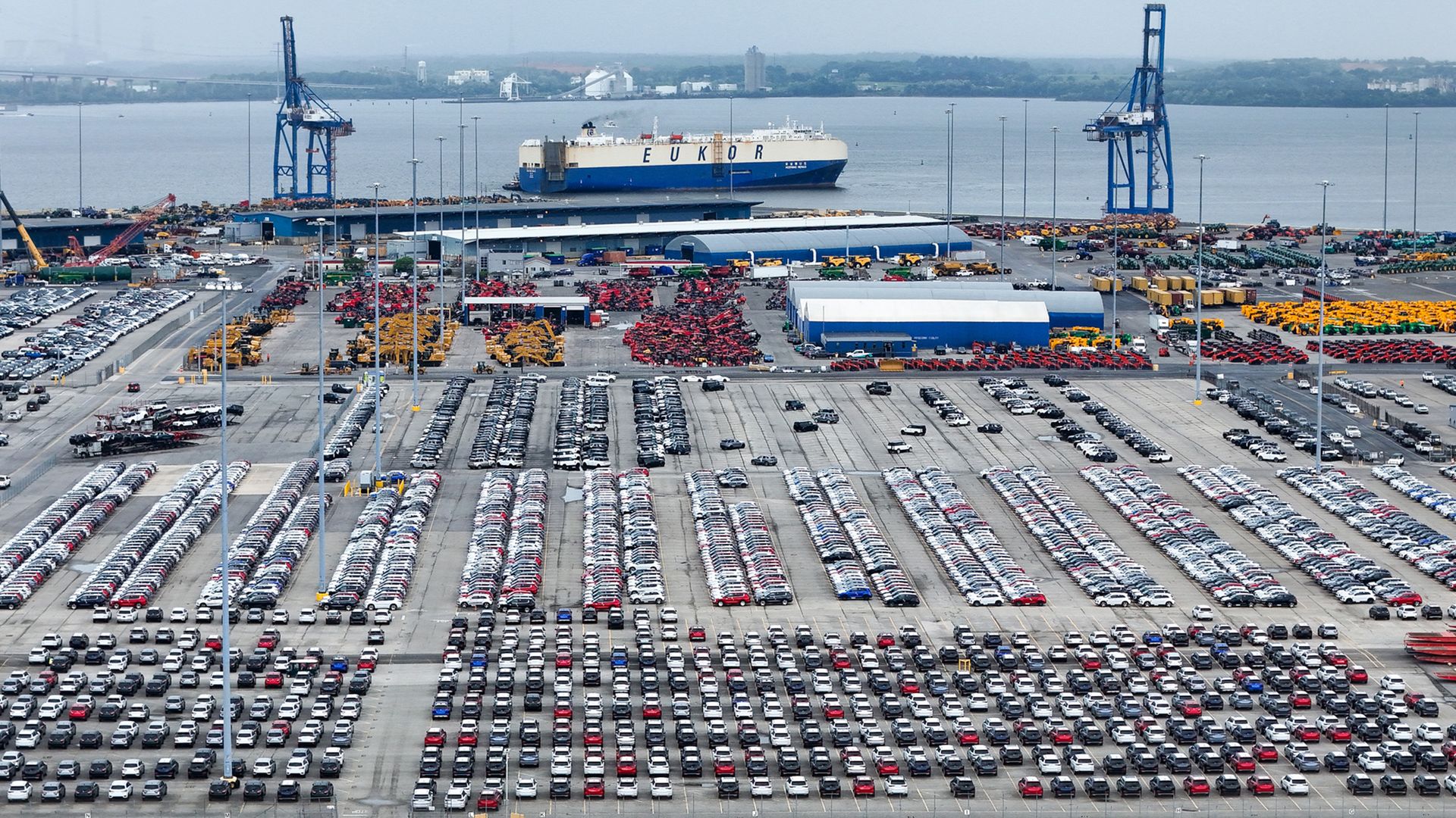 Aerial view of the Port of Baltimore, with rows of parked cars in the foreground, heavy machinery in yellow and red near blue warehouse buildings, and a large EUKOR cargo ship docked at the water's edge.