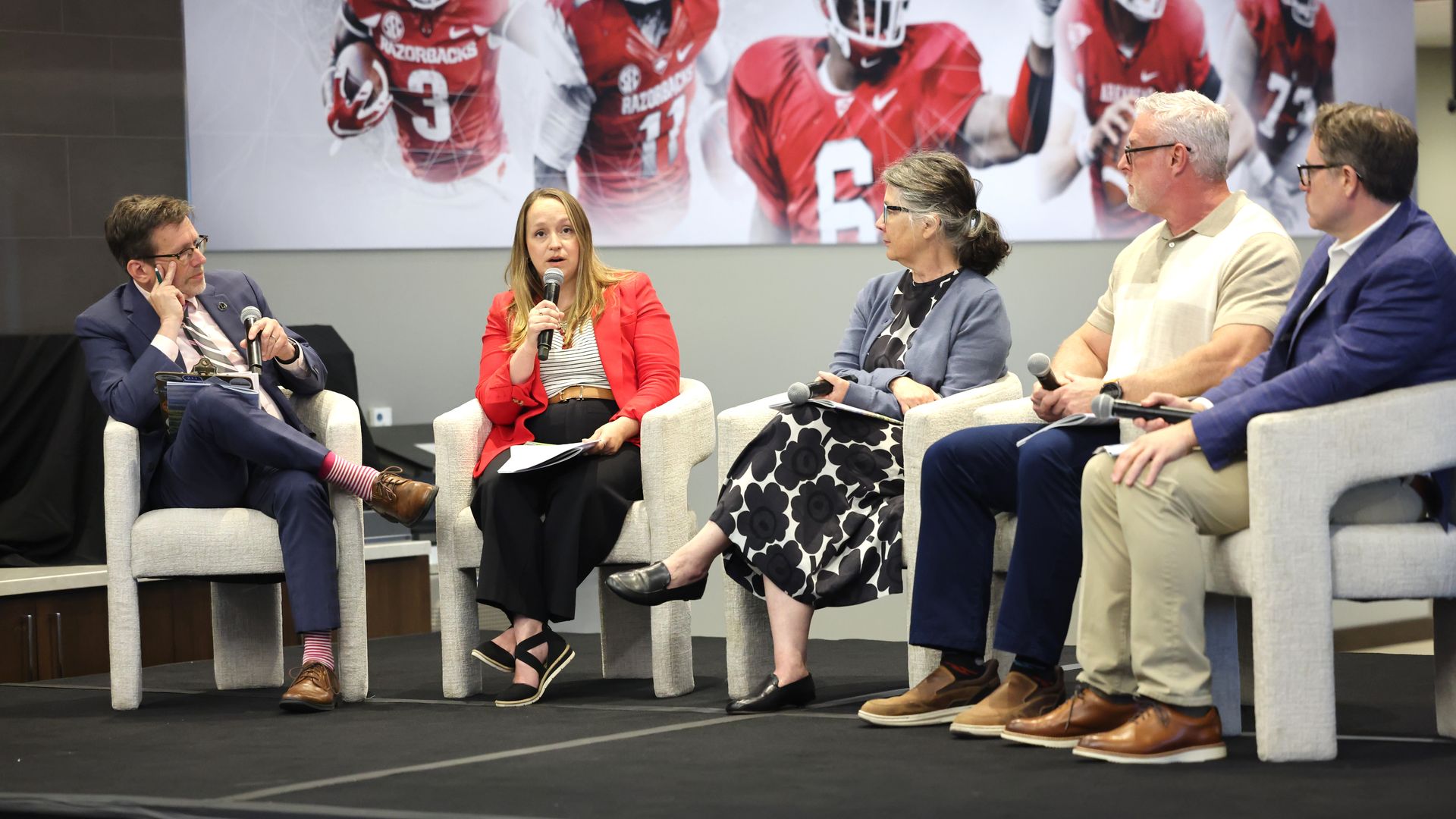 Five panelists sit in chairs on a stage; center woman in a red blazer speaks into a microphone, flanked by four others. A large red football-themed banner hangs behind them.