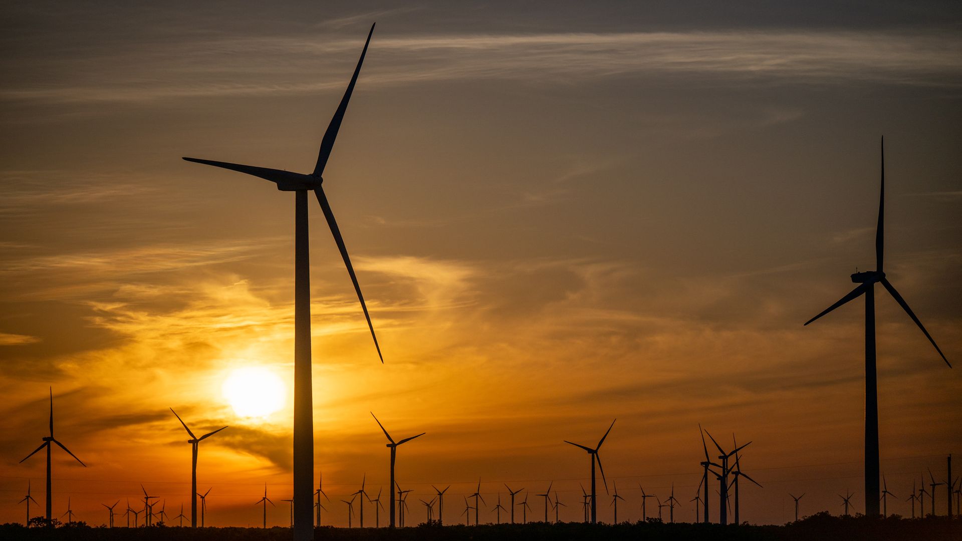 Wind turbines silhouetted against a golden orange sunset sky with scattered clouds over a vast field.