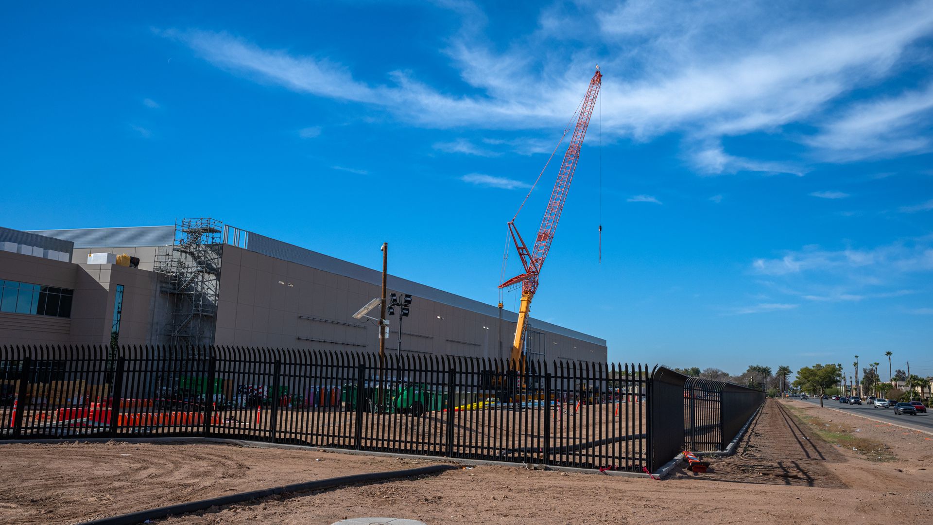 Construction site with a large building, black metal fence, and a red crane under a bright blue sky with wispy white clouds. Dirt ground and a road with cars visible to the right.