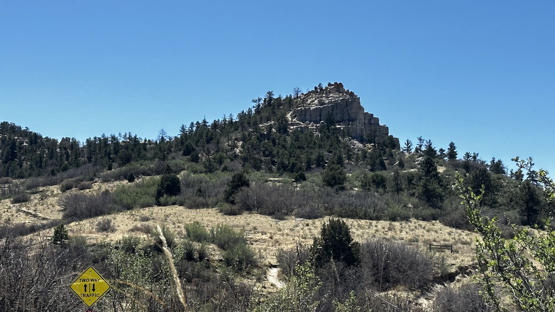 Rocky hill with a jagged summit and scattered pines rises above a dry grassy plain under a bright blue sky; a yellow "Two Way Traffic" sign is visible in the lower left.