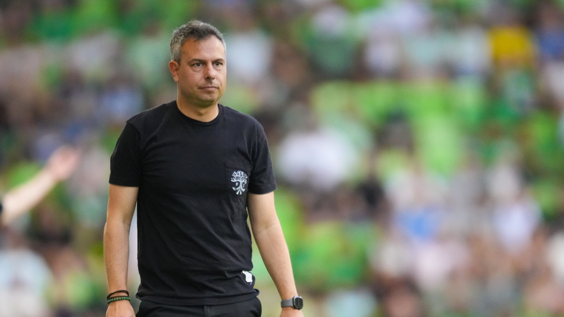 A man in a black T-shirt with a small white emblem on the chest stands on a sports field, with a blurred green crowd in the background.