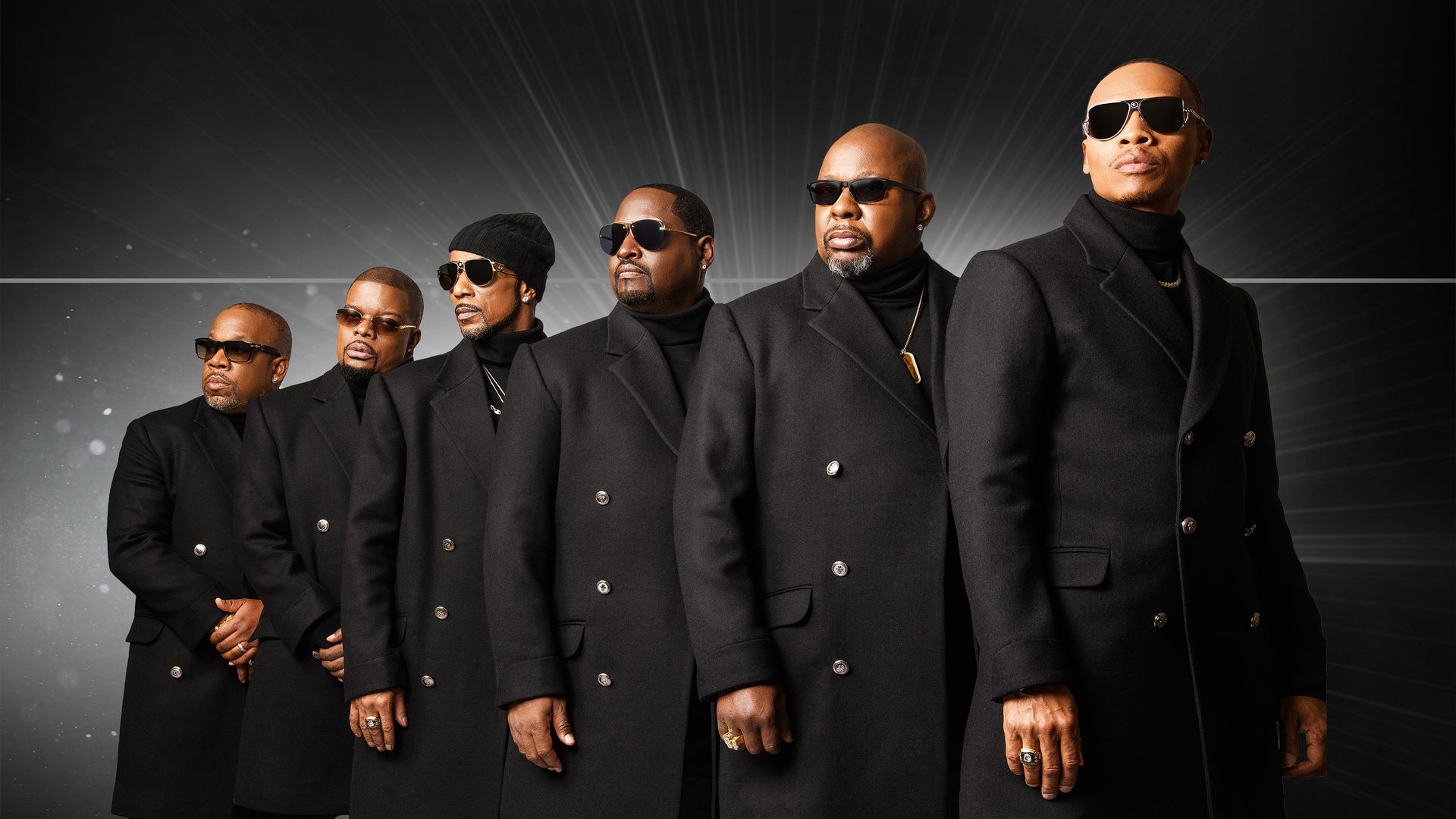 Six men in black overcoats and sunglasses stand in a diagonal line against a dark, radiant backdrop; one wears a knit cap; they pose confidently with rings and necklaces.