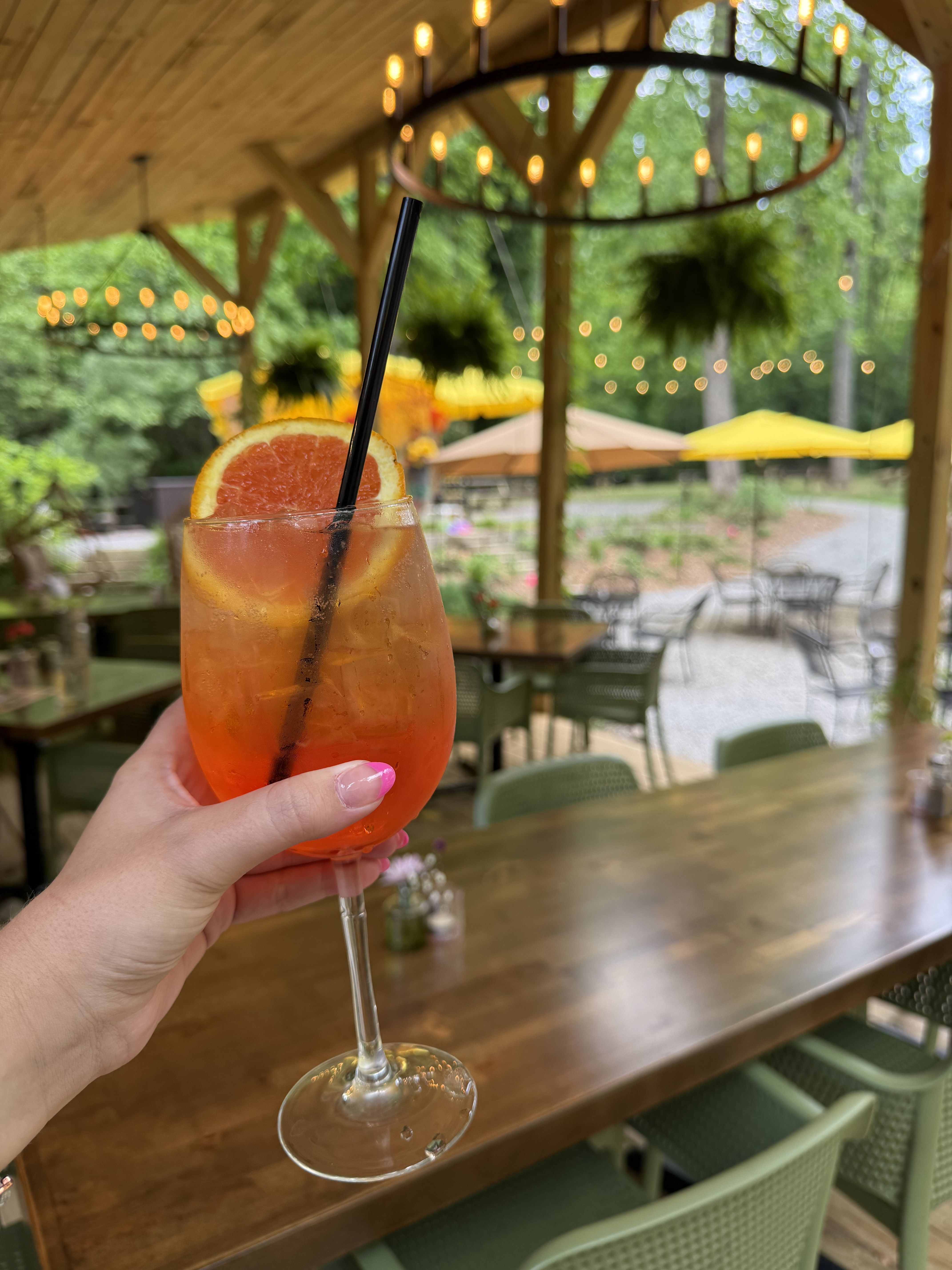Hand holds a tall stemmed glass of orange cocktail with a grapefruit slice garnish and black straw, set in an outdoor wooden cafe with string lights and yellow umbrellas, blurred background.