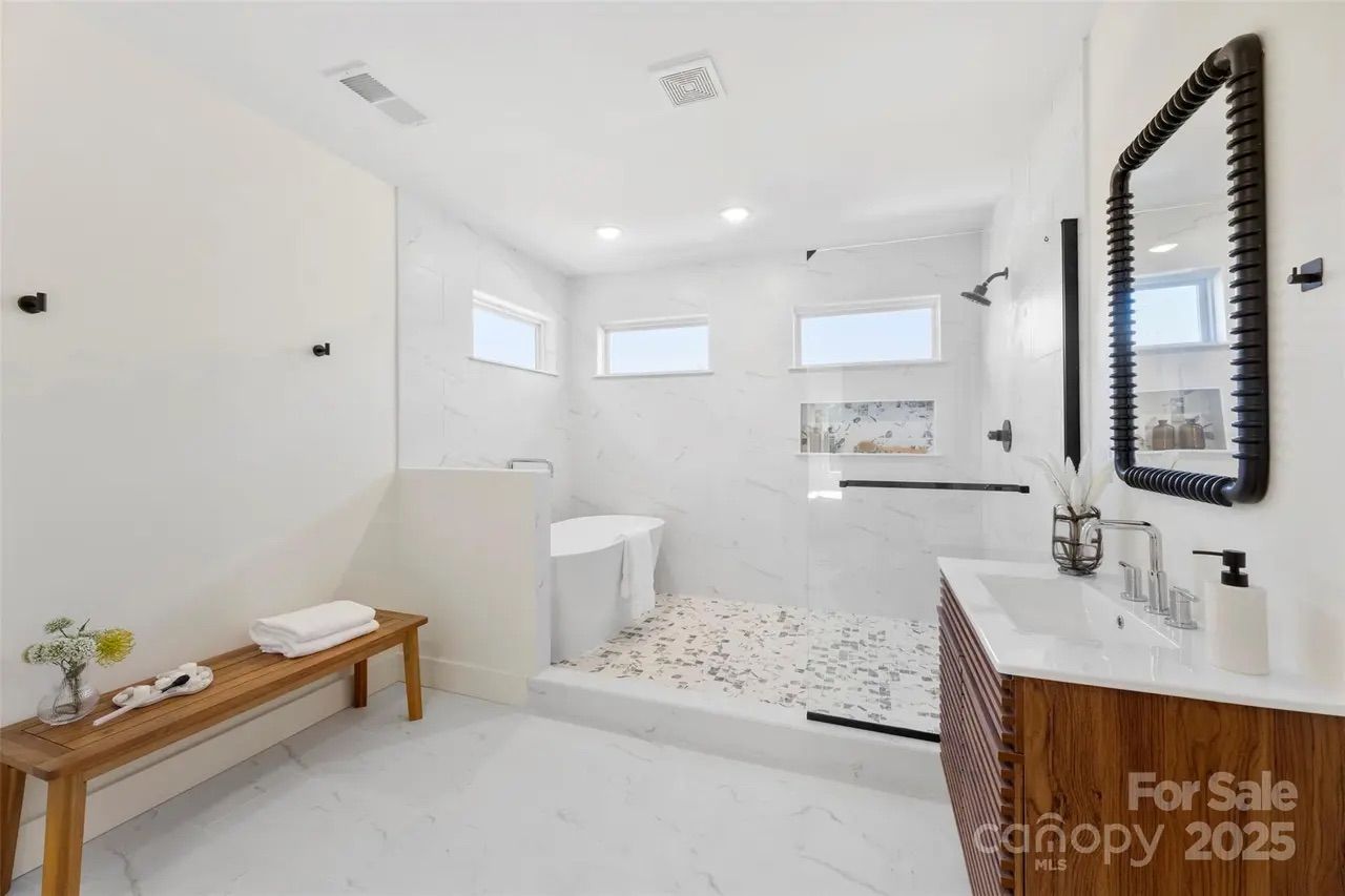 Modern white bathroom with a freestanding tub, walk-in shower with mosaic tile floor, wooden vanity with black mirror, and a wooden bench with towels and flowers on it.