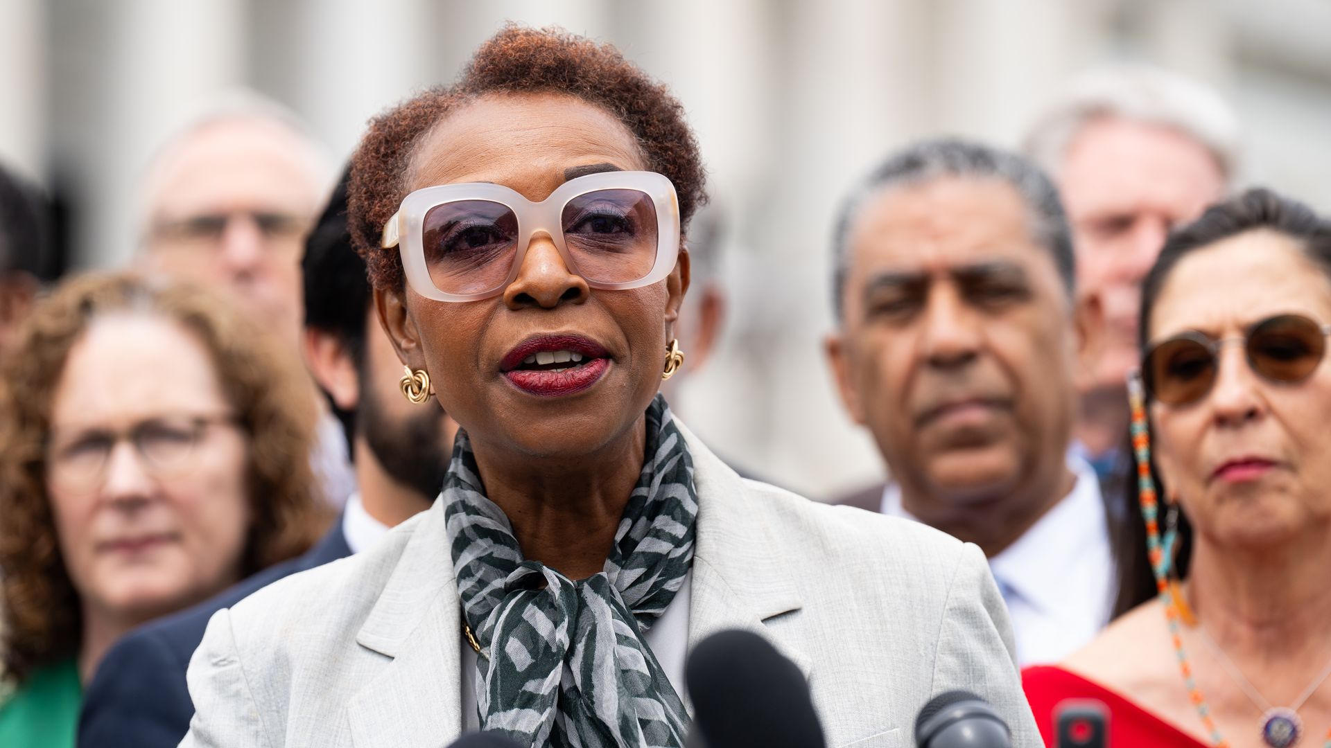 Rep. Yvette Clarke, wearing a white blazer and sunglasses, speaks into a microphone in front of colleagues outside the U.S. Capitol.