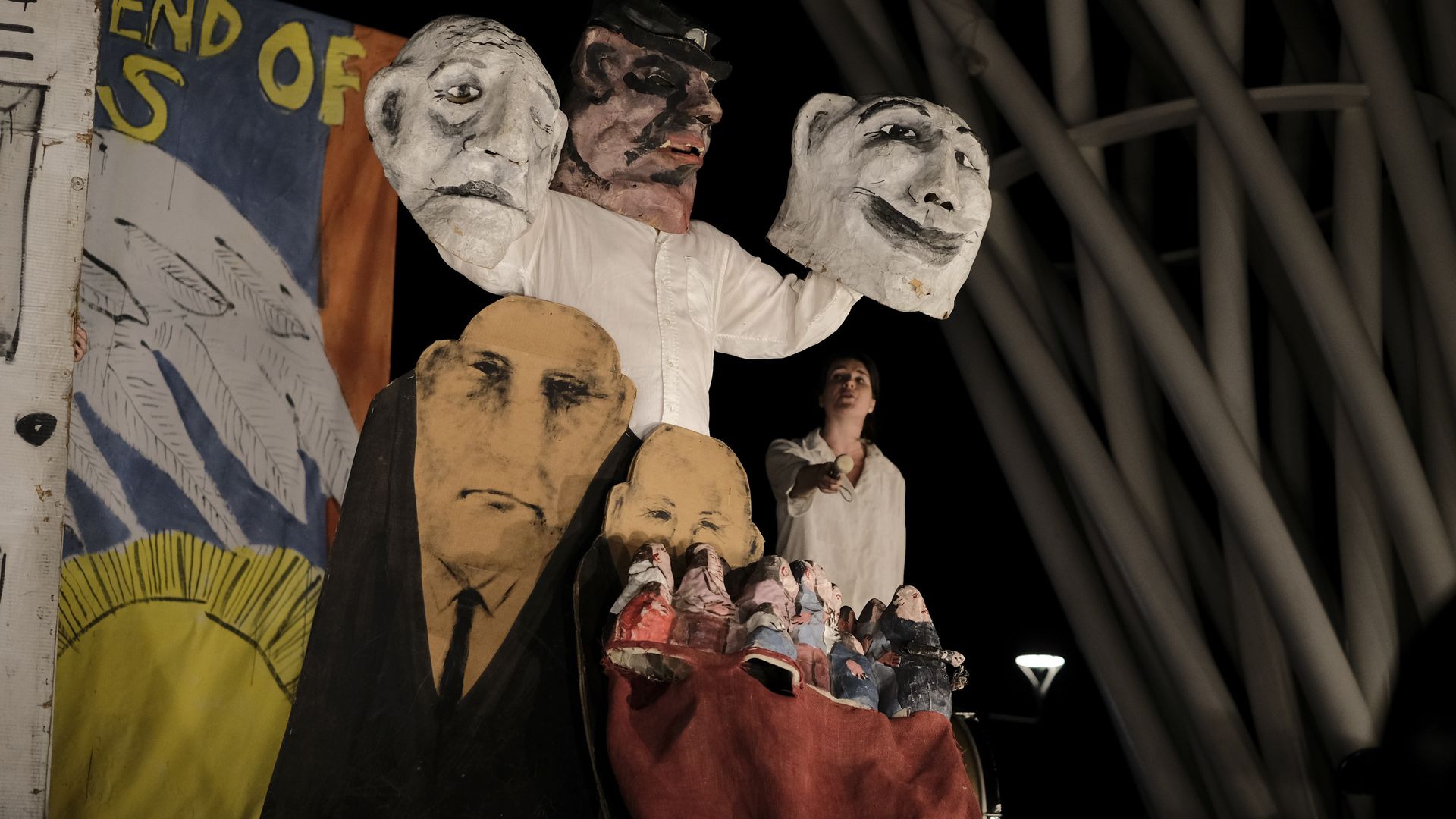 Nighttime puppet show featuring oversized paper-mâché heads on a raised platform. A performer in white holds white-faced masks and a pink-toned head, with a painted backdrop and smaller faces below.