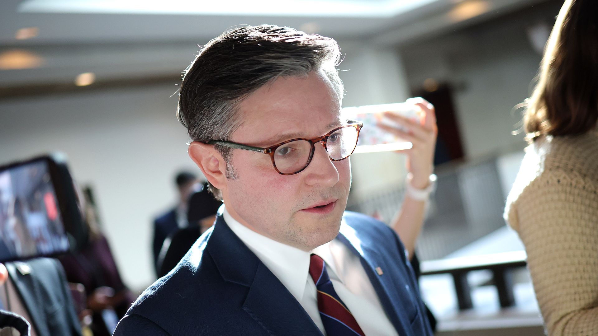 Man with gray-streaked hair wearing glasses, navy blue suit, white shirt, and red-striped tie indoors with blurred people and bright lighting in the background.