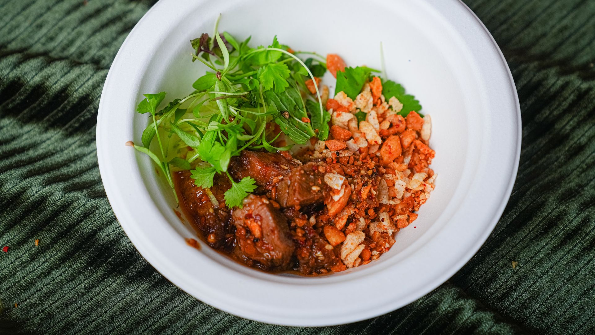 White bowl containing braised meat in tomato sauce, topped with fresh greens and microgreens, plus orange crumbly topping, set on a dark green textured cloth.