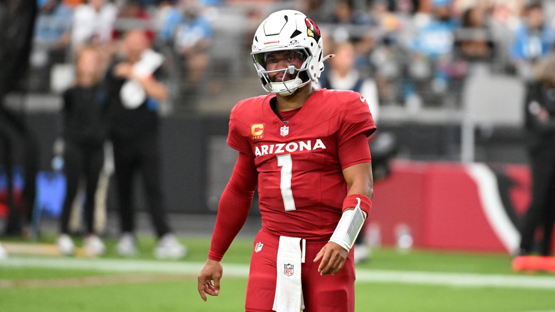 A football player in a red uniform and white helmet, with the word "Arizona" and the number "1" in white on the front of the jersey, stands on the sideline of a football field, with people standing in the background. 