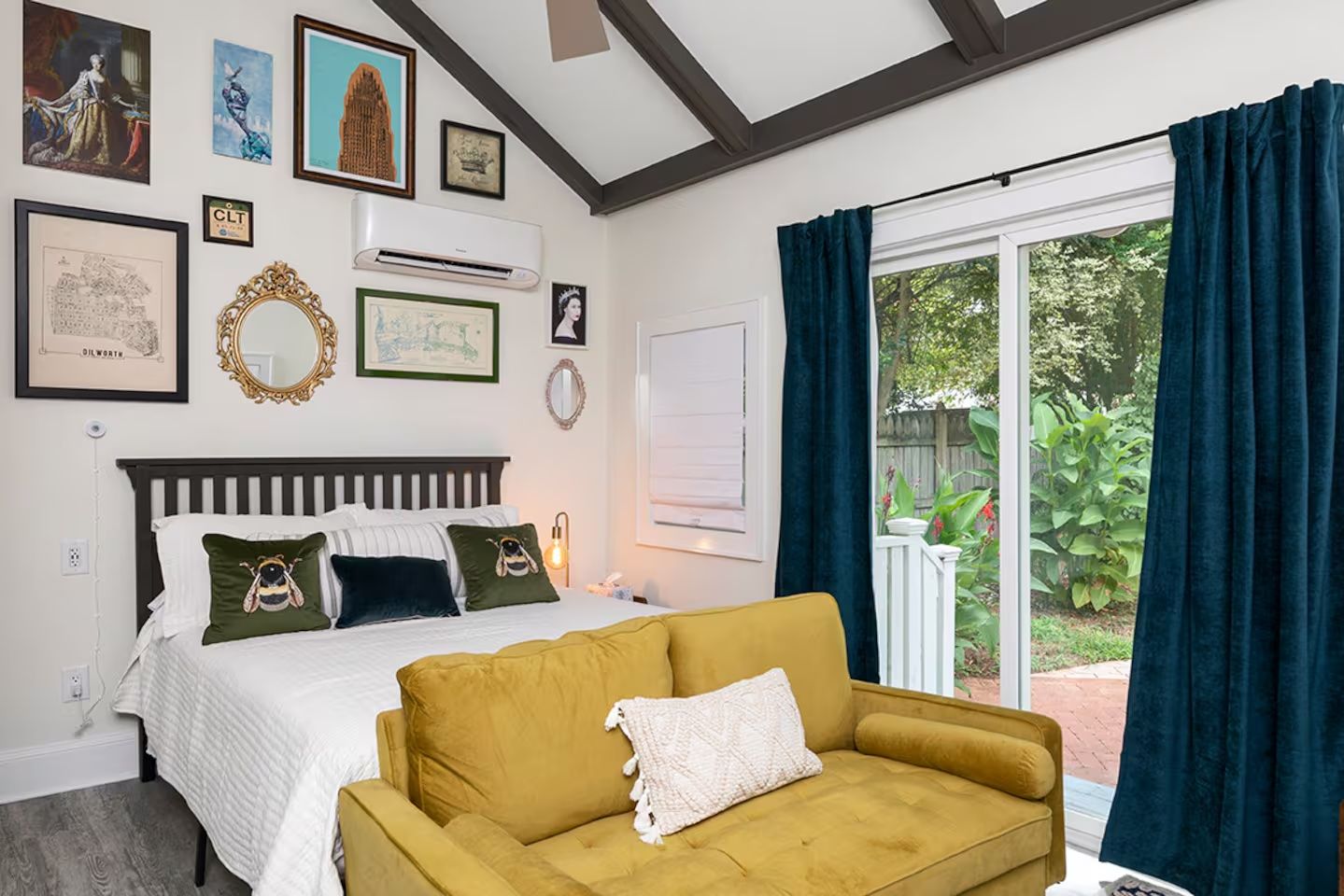 Bedroom with white walls and dark ceiling beams, bed with white bedding and bee-themed green pillows, mustard yellow sofa, dark blue curtains, sliding glass door to garden, and framed artwork on walls.