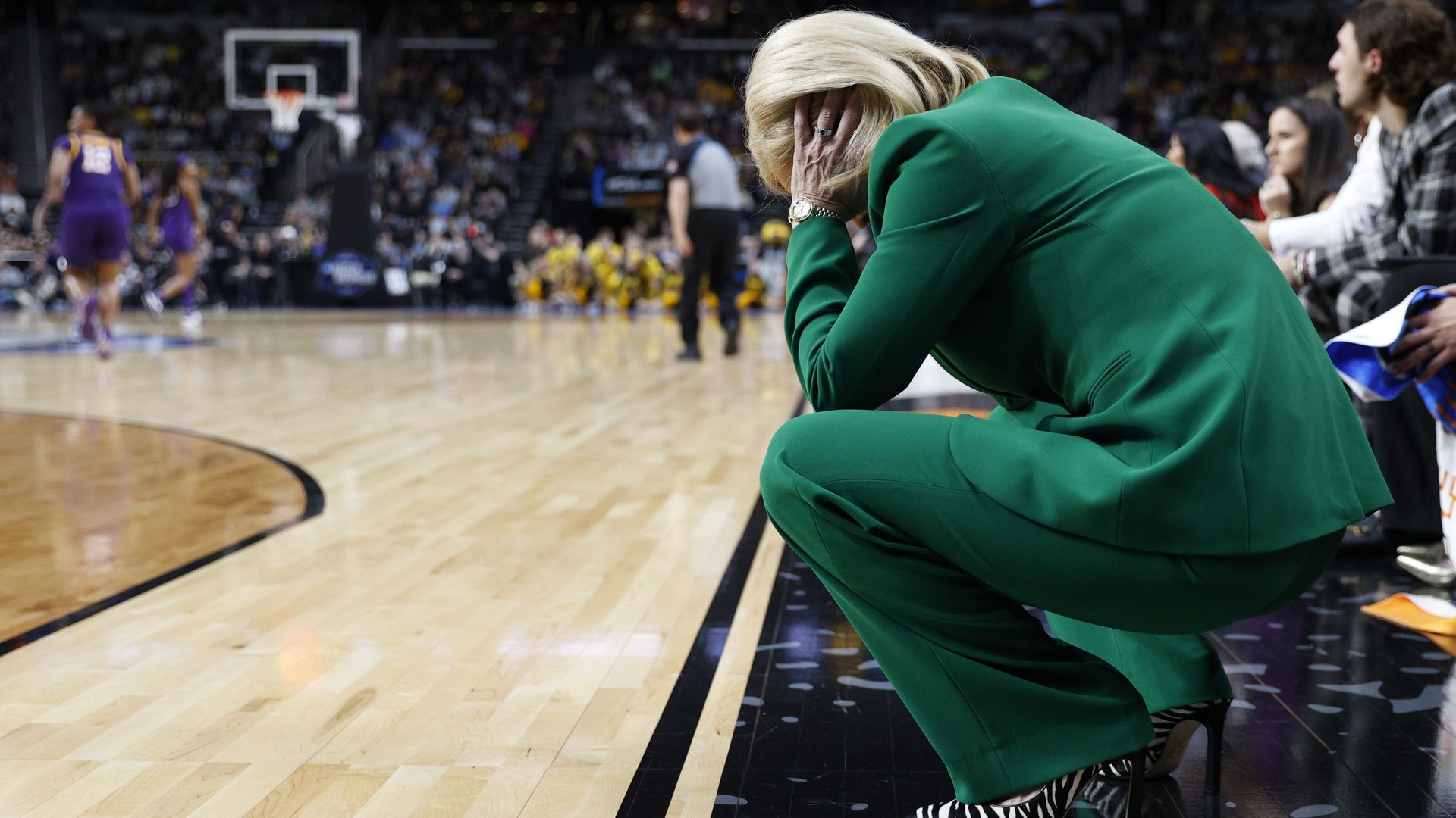 Kim Mulkey kneels on the sideline with her head in her hands as her team plays on the court.
