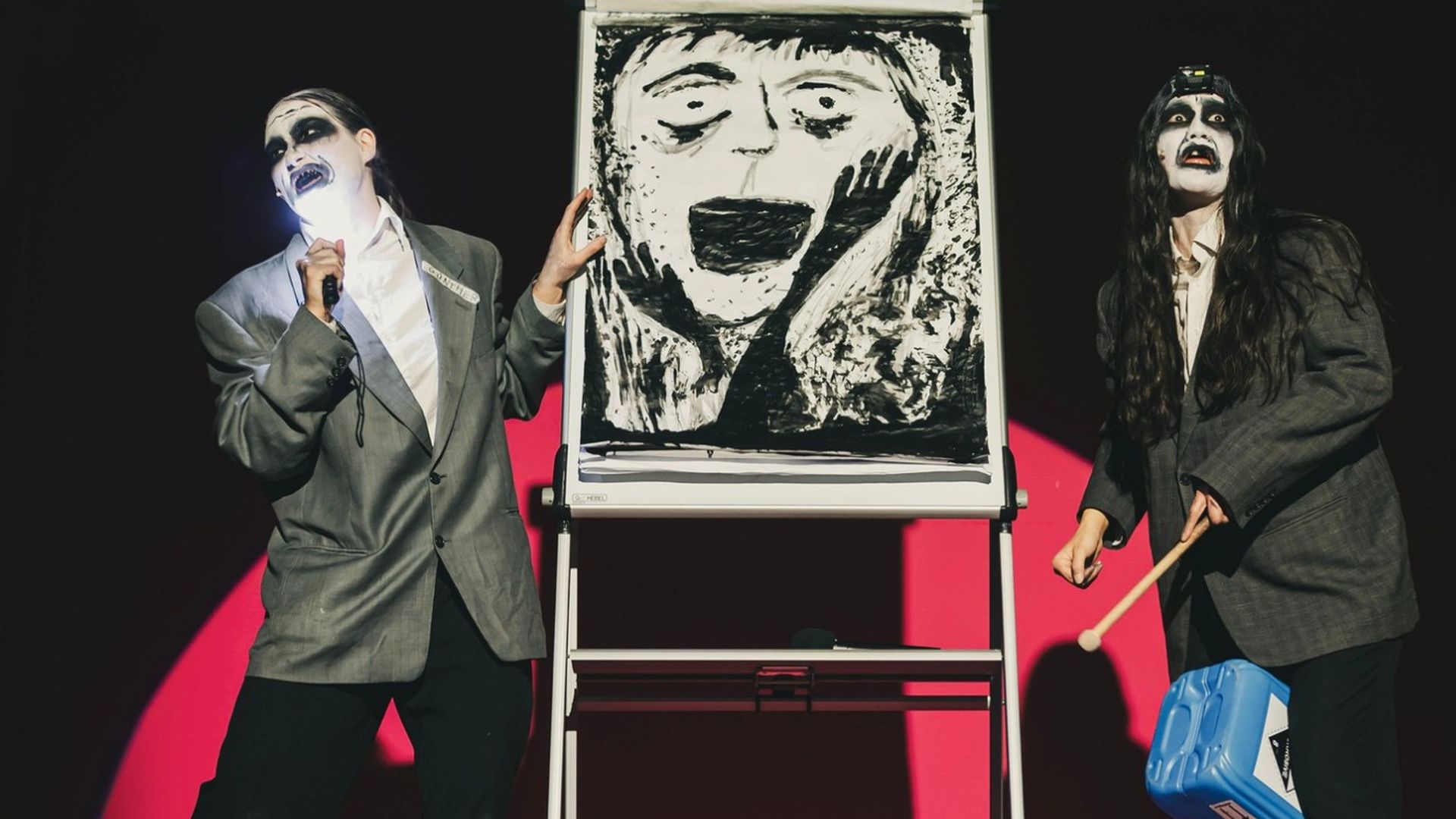 Two performers in gray suits and white-face makeup perform on stage. Left singer with microphone gestures to a large black-and-white scream drawing on a flip chart; right performer holds a blue container.