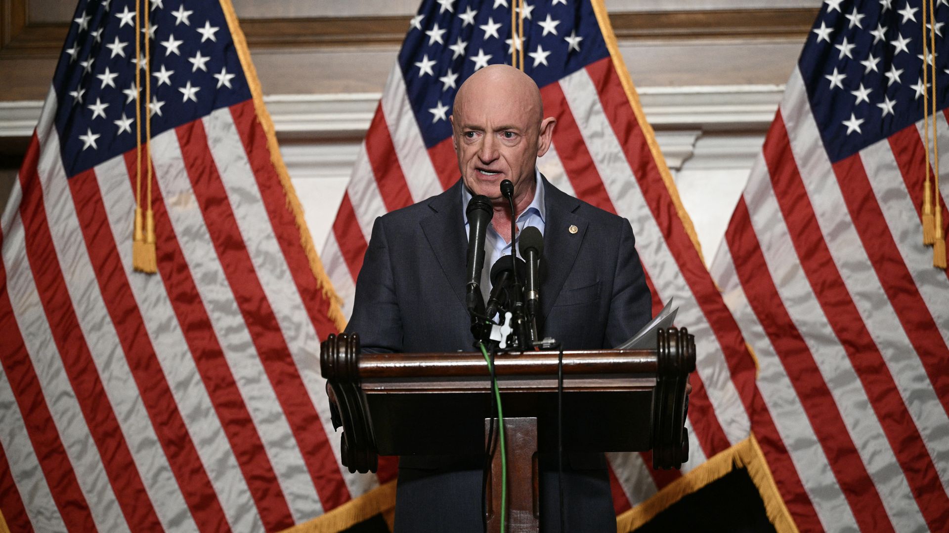 Bald man in a dark suit speaking at a wooden podium with multiple microphones, with three American flags with gold fringe in the background.