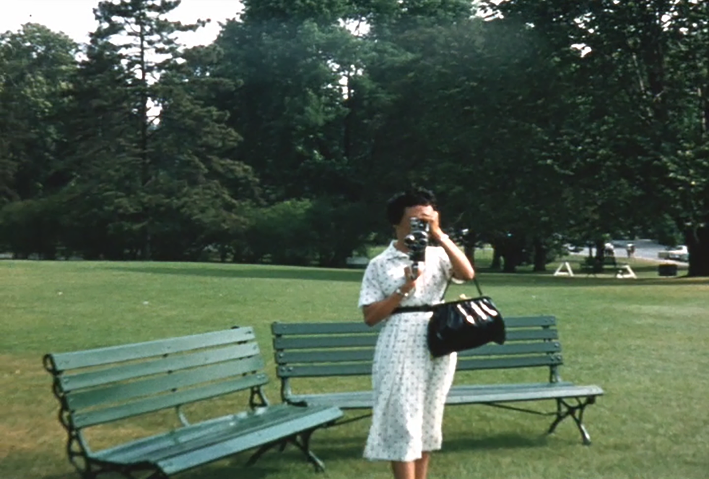 A still of a woman in a white dress holding an old video camera.