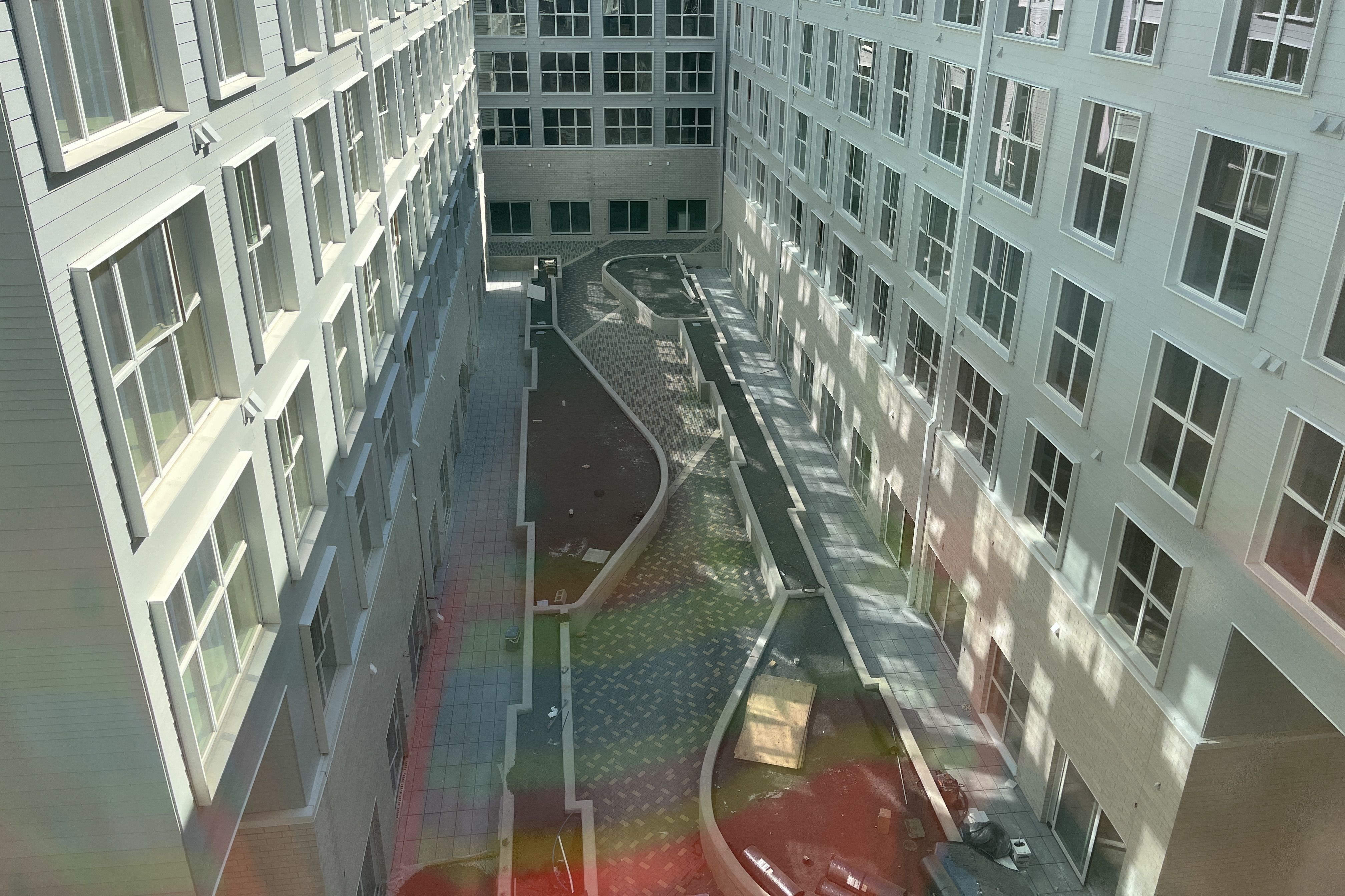 Bird's-eye view of a modern apartment complex courtyard: pale blue building facades with many windows flank a central, open, tiled atrium featuring curved stair rails and shaded walkways.