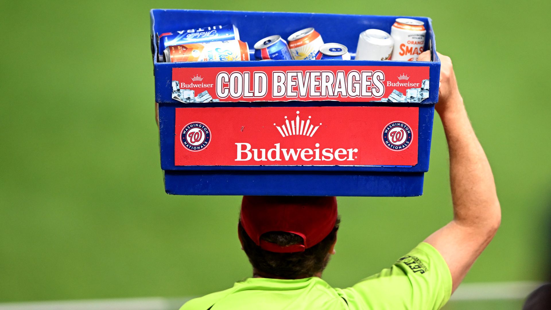 Back view of a person wearing a bright lime shirt and red cap carrying a blue crate labeled "COLD BEVERAGES" with Budweiser branding, filled with assorted soda cans, against a green blurred background.