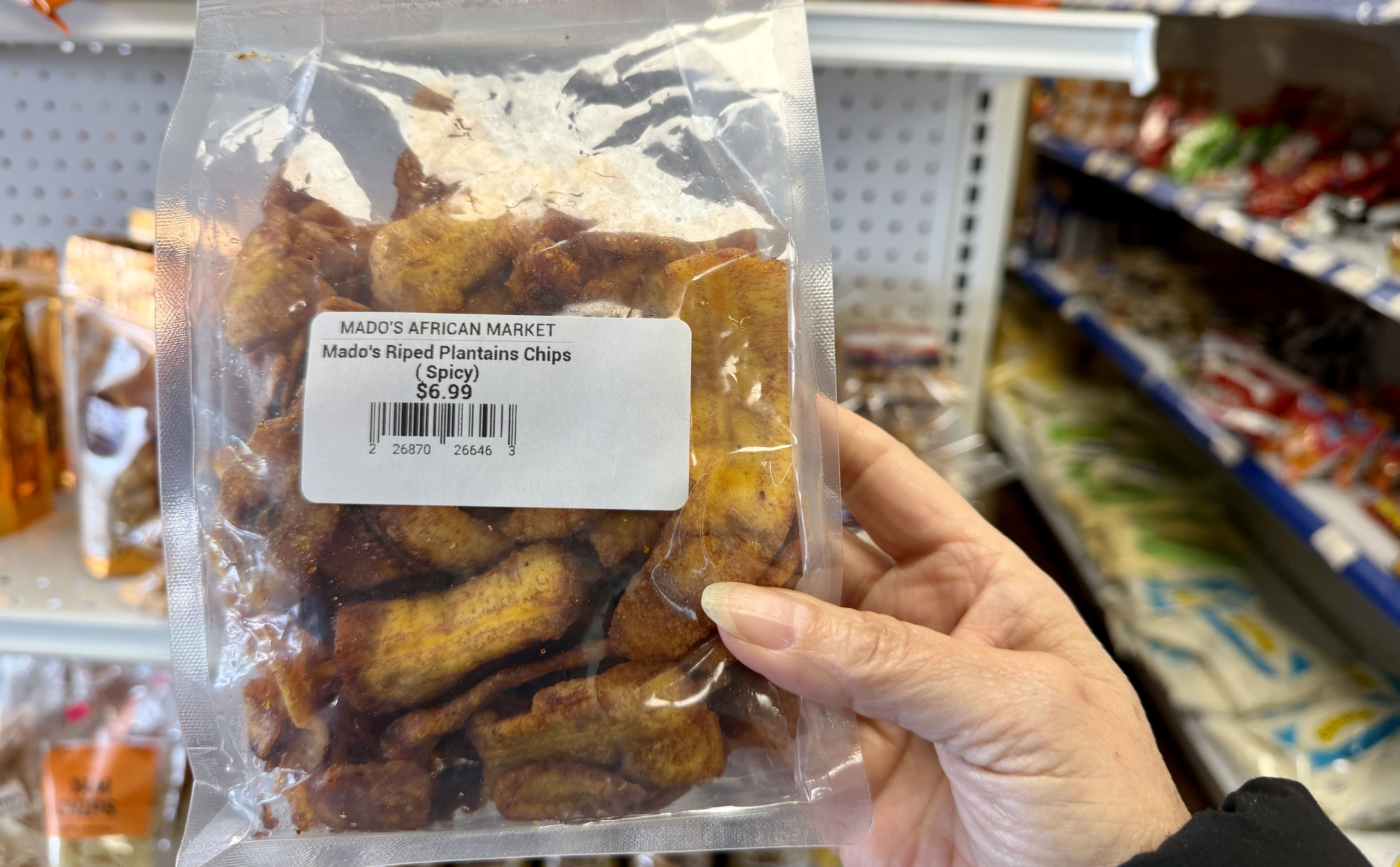 A hand holds a clear bag of Mado's African Market spicy plantain chips in a grocery store; golden-brown chips visible through plastic, label shows $6.99.