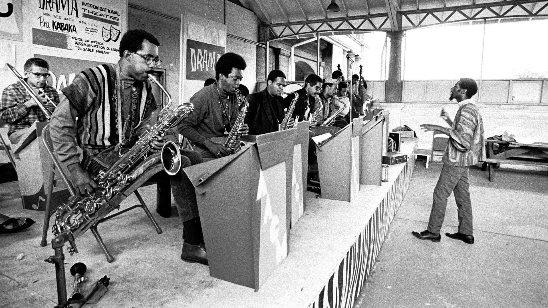Black-and-white scene of a band on an outdoor stage; musicians with saxophones and brass sit at stands, facing a conductor in a striped shirt, with "DRAMA" posters on the wall.