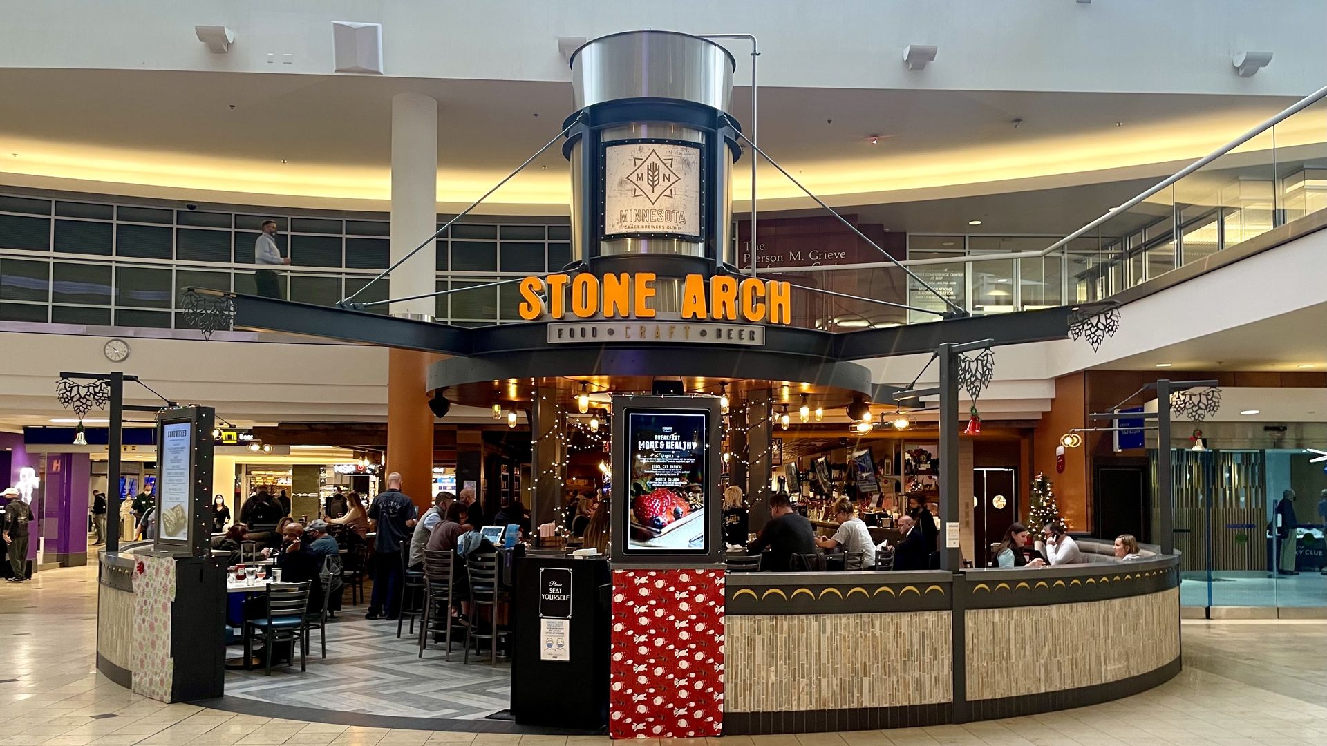 A large restaurant in the center of the airport, with people eating at round tables. An orange sign reads Stone Arch.