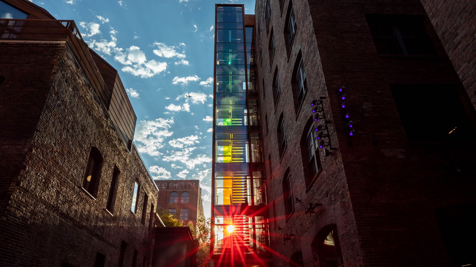 A photo of two brick buildings with a rainbow colored glass structure with bright light coming through in between the buildings.