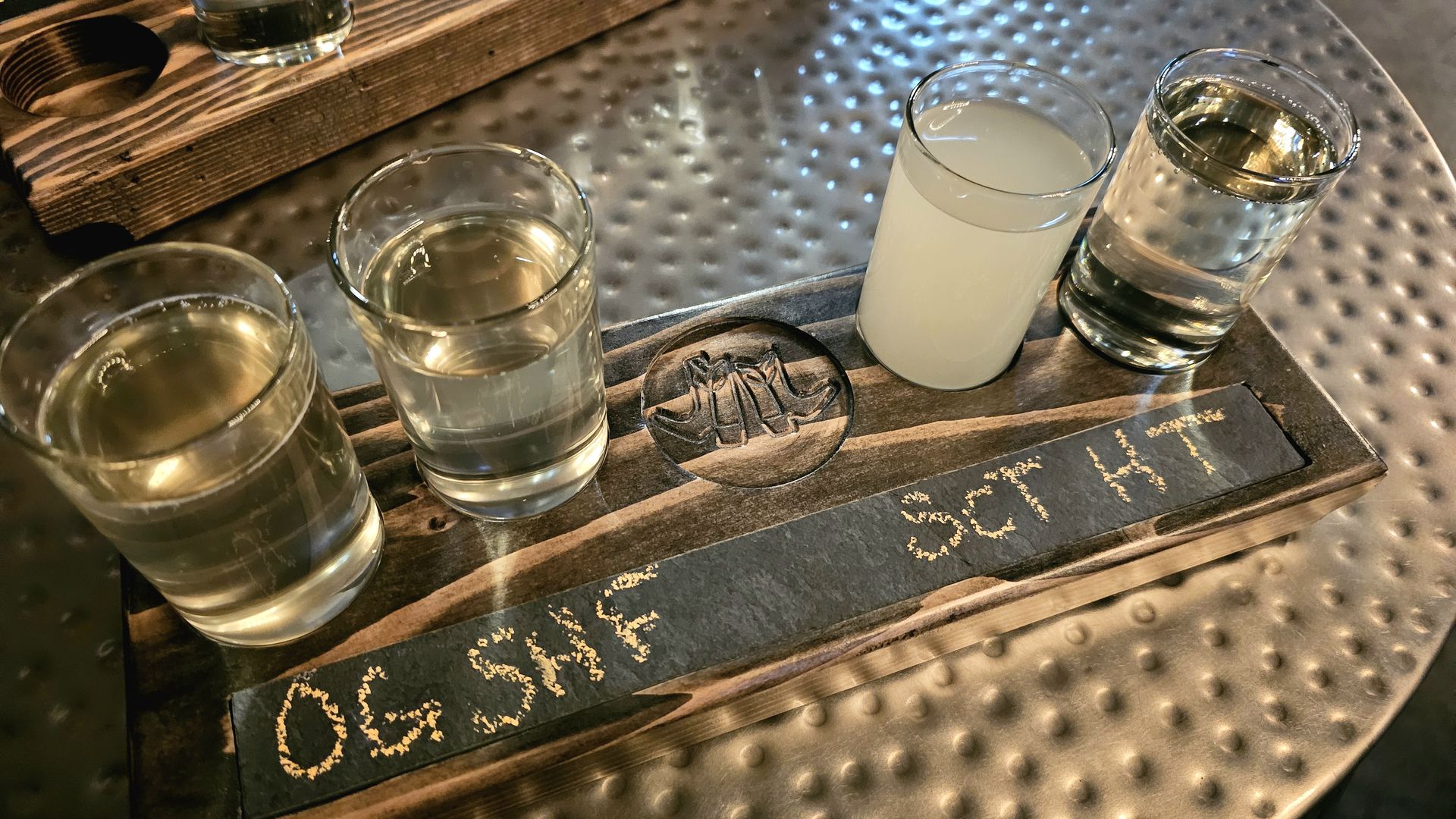 Four-glass beer flight on a wooden tray, with amber, pale yellow, cloudy white, and clear drinks.