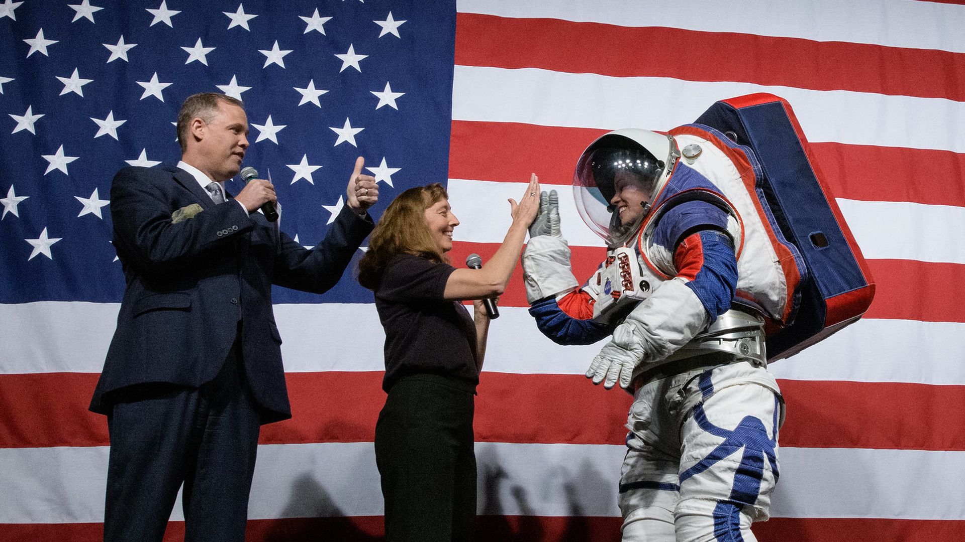 A person wears a spacesuit next to two other people in front of an American flag