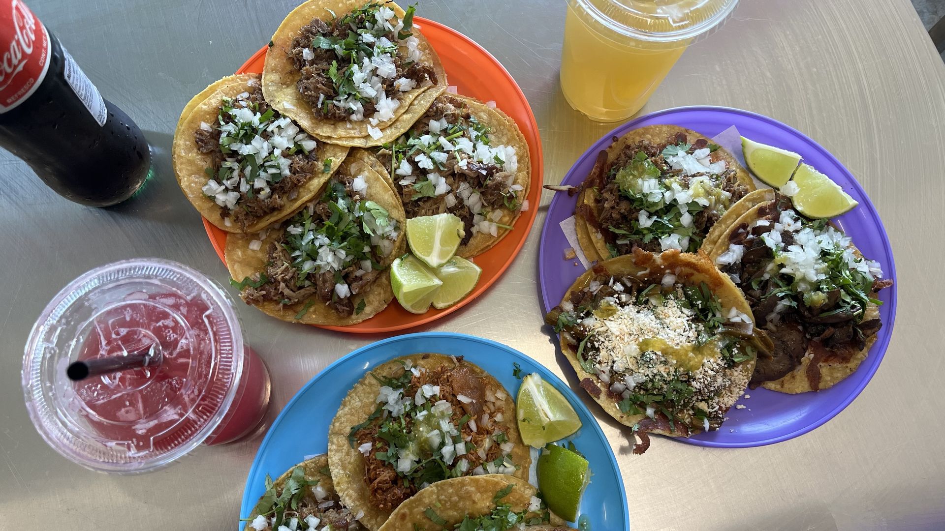 Three colorful plates with tacos topped with chopped onions, cilantro, and lime wedges on a metal table. Drinks include a Coca-Cola bottle, a pink drink with a straw, and a yellow drink in plastic cups.