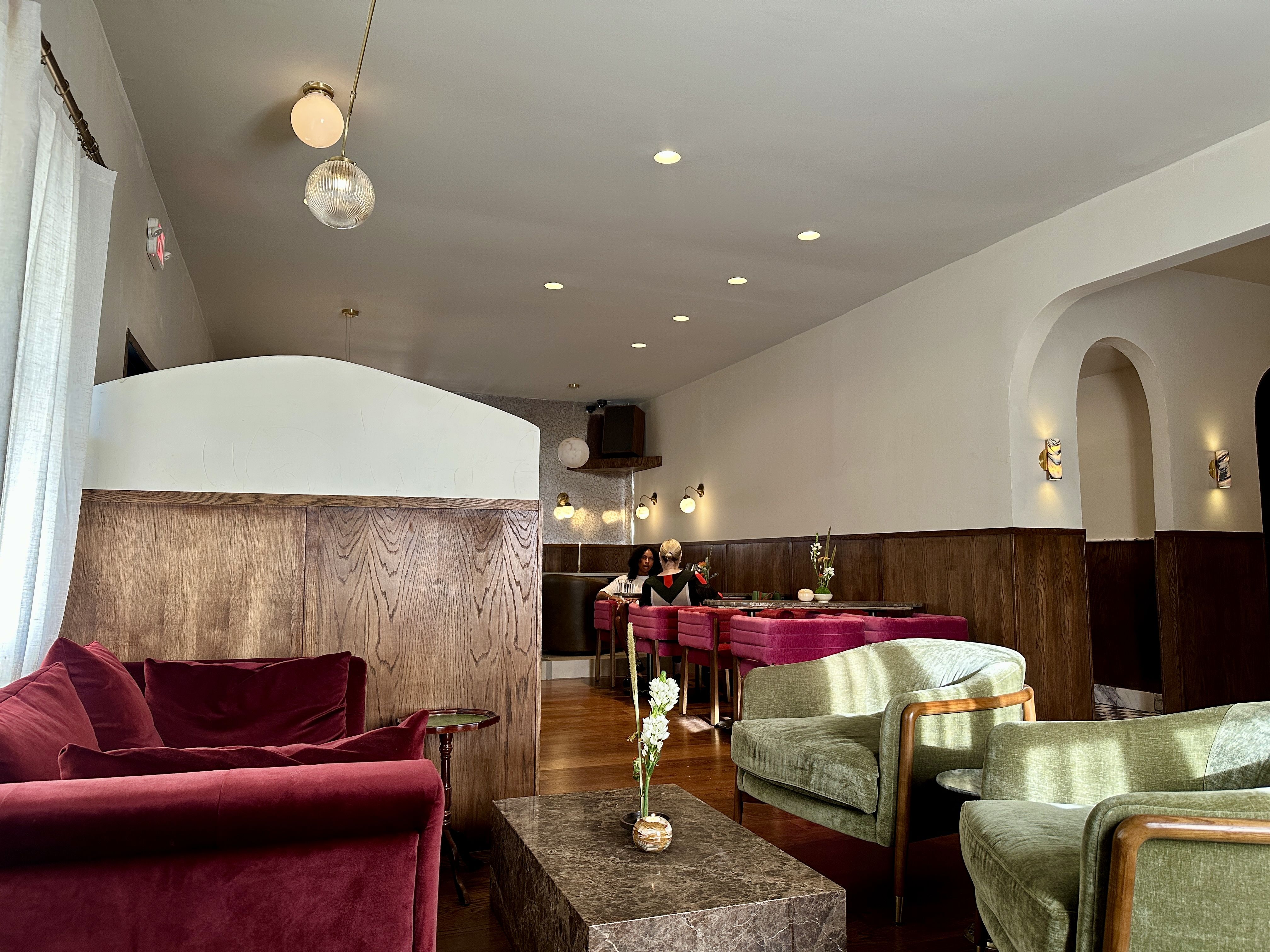 A stylish lounge with burgundy velvet sofas and green chairs, wood-paneled walls, marble coffee table, and warm pendant lights. Arched doorway on the right, curtains left, dining area in back. Huckleberry Noda