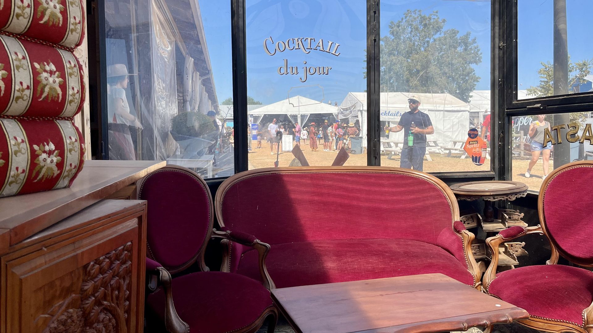 Interior of a glass-enclosed seating area with red velvet vintage sofa and chairs around a wooden table, overlooking an outdoor event with people, tents, and a sign reading "Cocktail du jour".